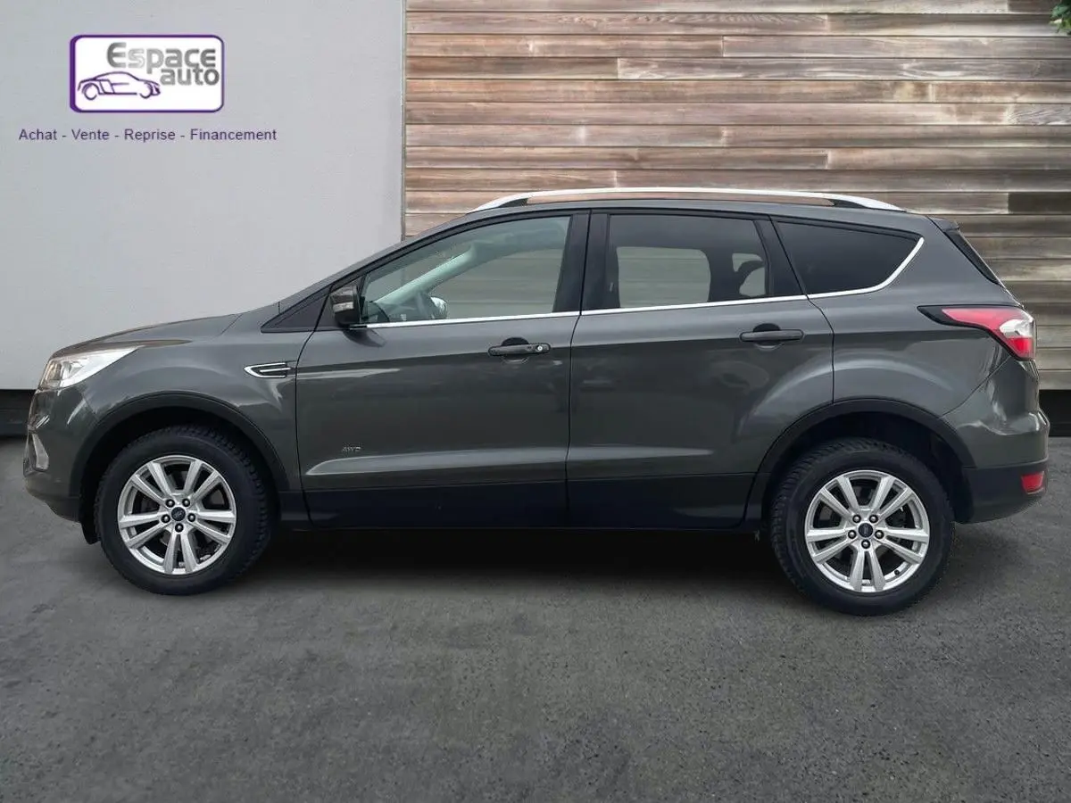 Profil côté gauche d'un Ford Kuga gris 2017 avec jantes alliage et barres de toit, garé devant un mur en bois.