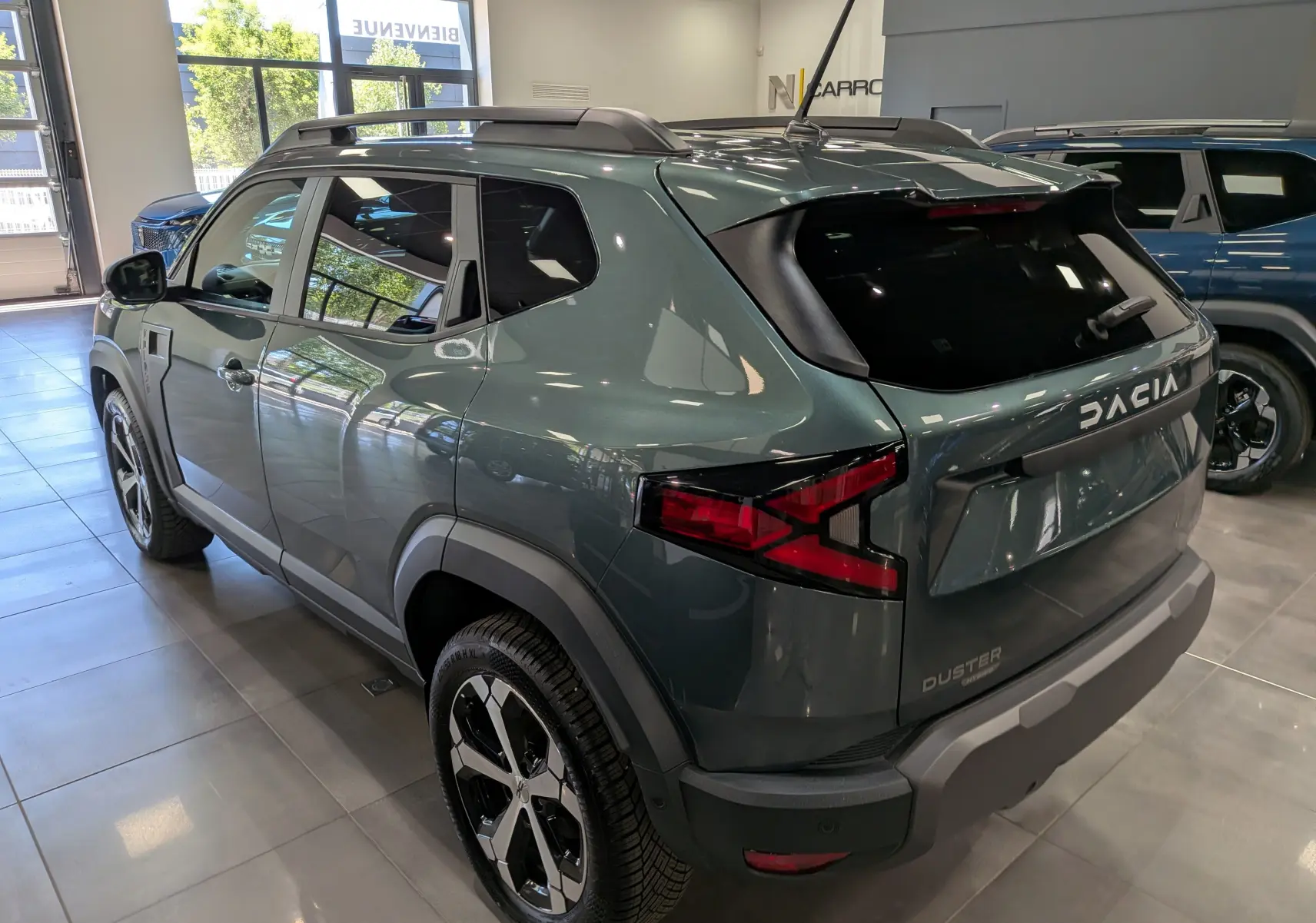 Vue 3/4 arrière droite d'un Dacia Duster Hybrid-G 150 CH 4x4 Journey vert cèdre avec jantes biton et barres de toit.