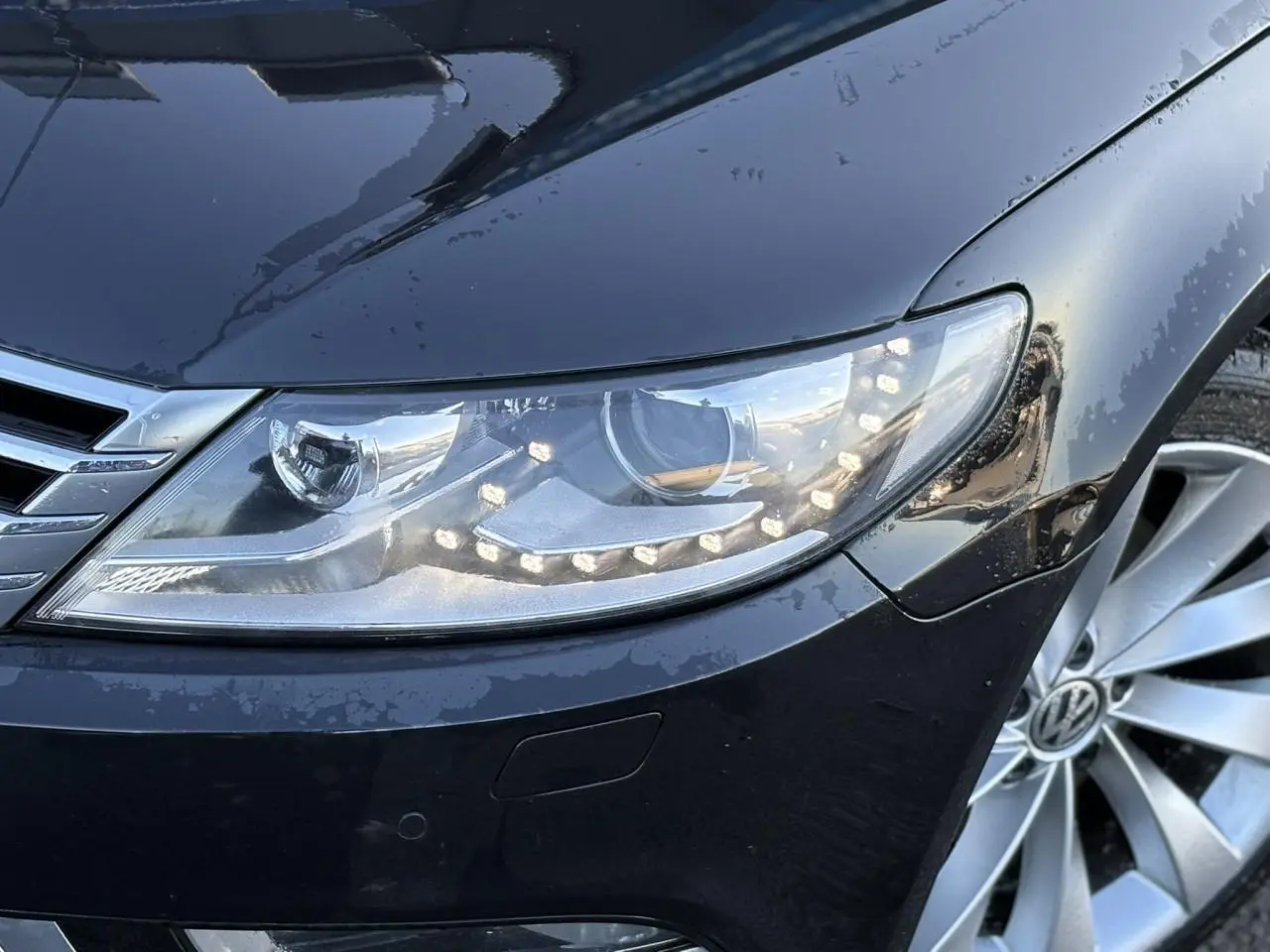 Gros plan sur le phare avant droit et la jante alliage 18'' argentée d'une Volkswagen Passat CC Deep Black 2015.