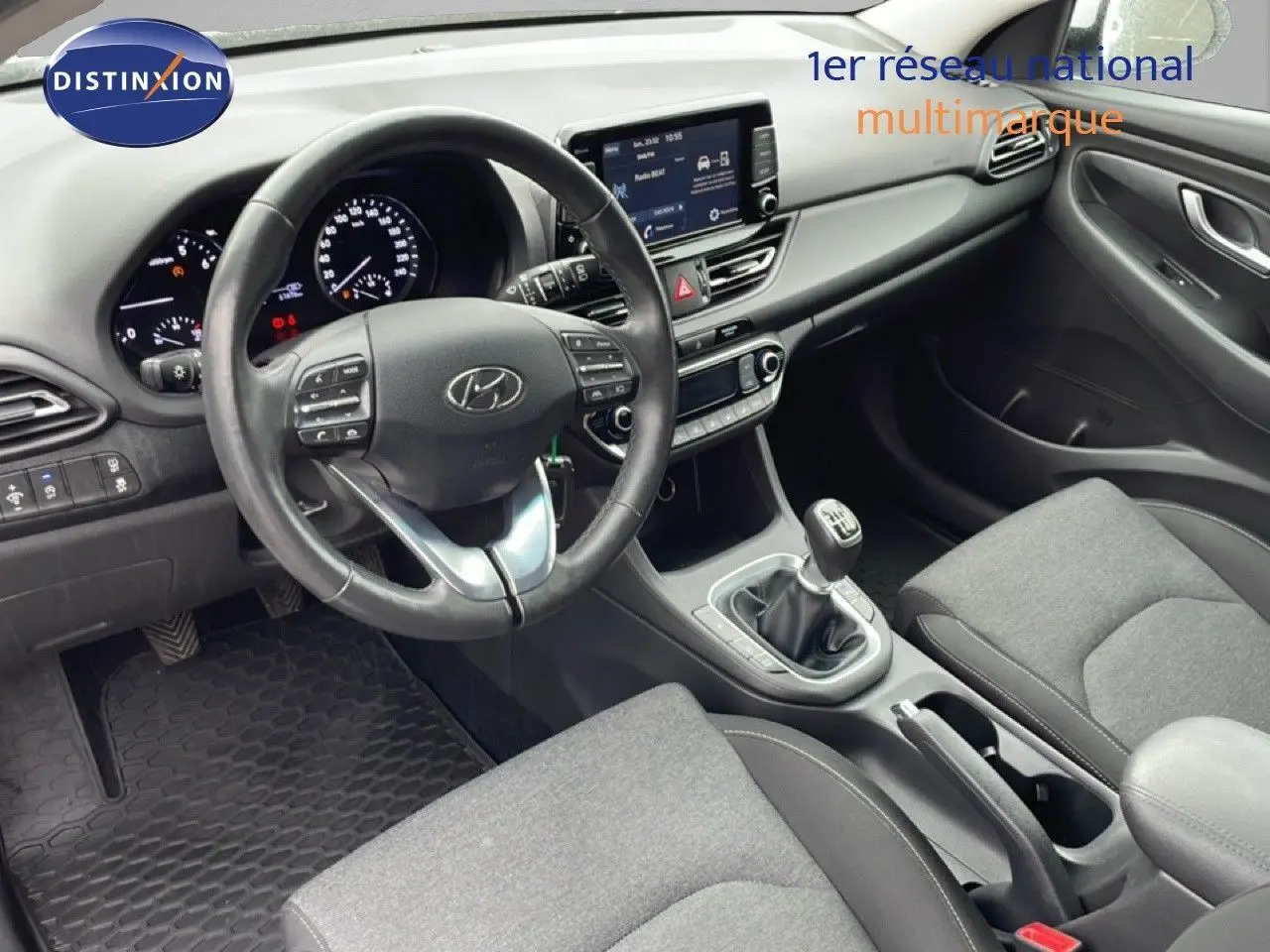 Vue intérieure de face du poste de conduite de la Hyundai i30 SW blanche, volant multifonction et boîte manuelle visibles.