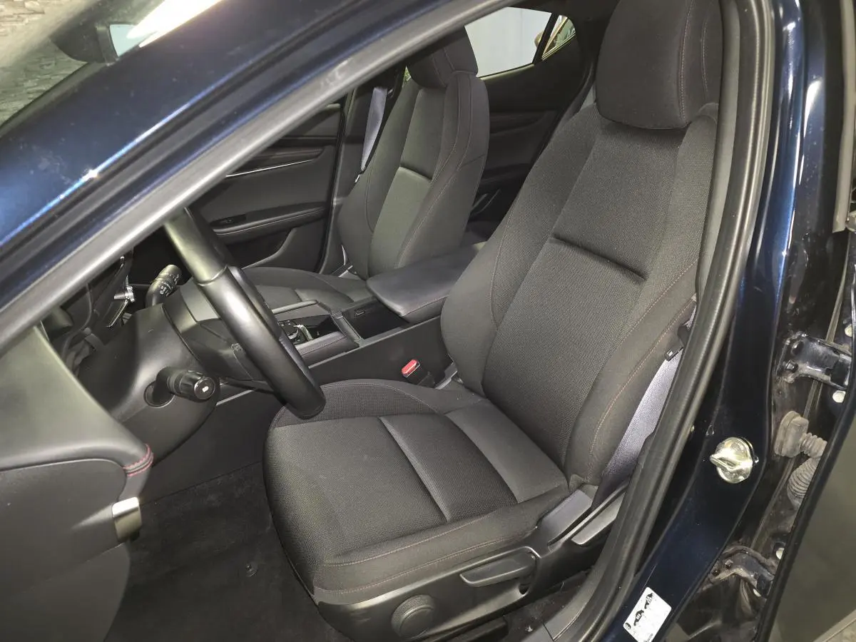 Vue intérieure côté conducteur de la Mazda3 2024 bleu, sièges tissu noir avec surpiqûres rouges et accoudoir central.
