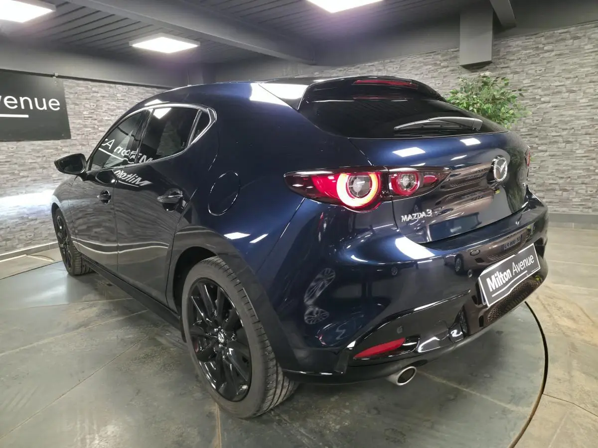 Vue 3/4 arrière droite d'une Mazda3 2024 bleu métallisé avec jantes noires et feux arrière LED circulaires allumés.