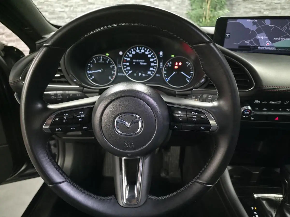 Vue intérieure centrée sur le volant cuir noir de la Mazda3 2024 avec tableau de bord et écran de navigation visibles.