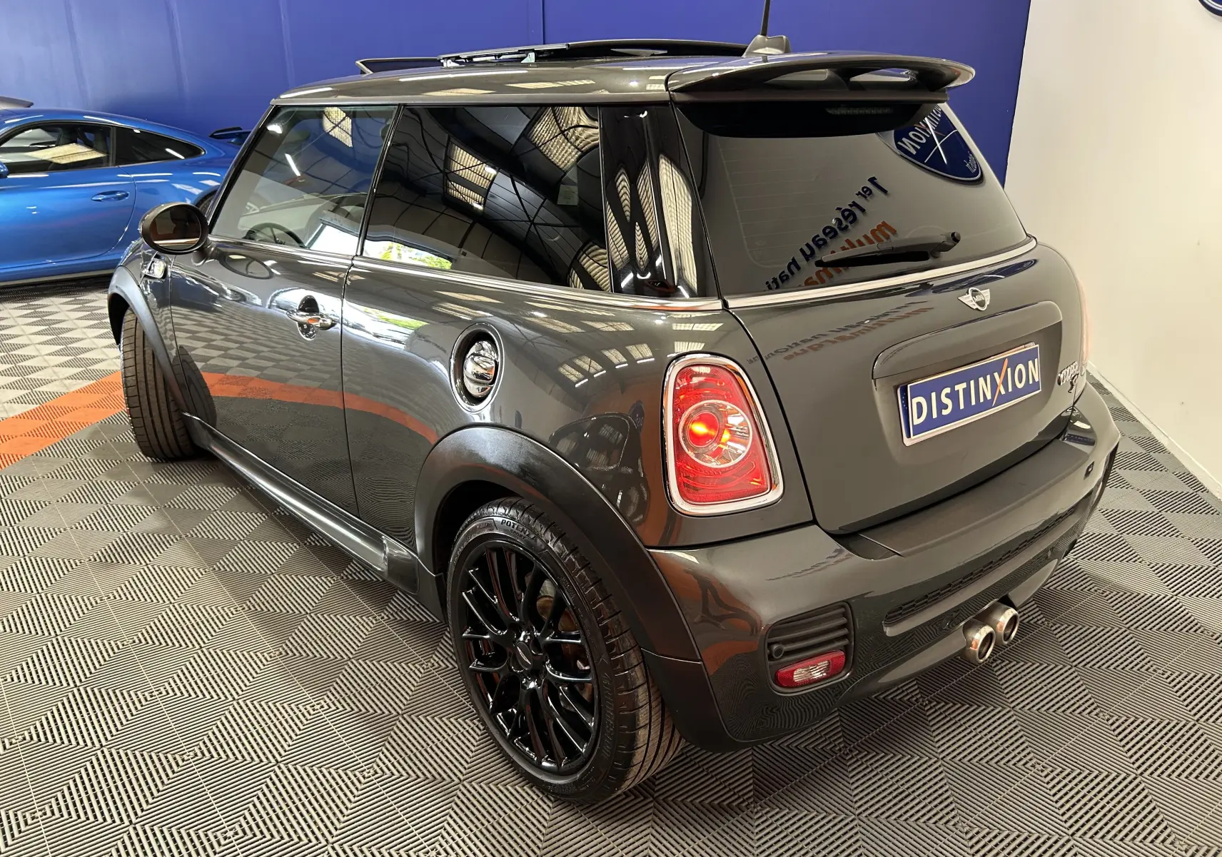 Mini Cooper gris vue 3/4 arrière droit, jantes noires brillantes et feux arrière allumés en intérieur showroom.