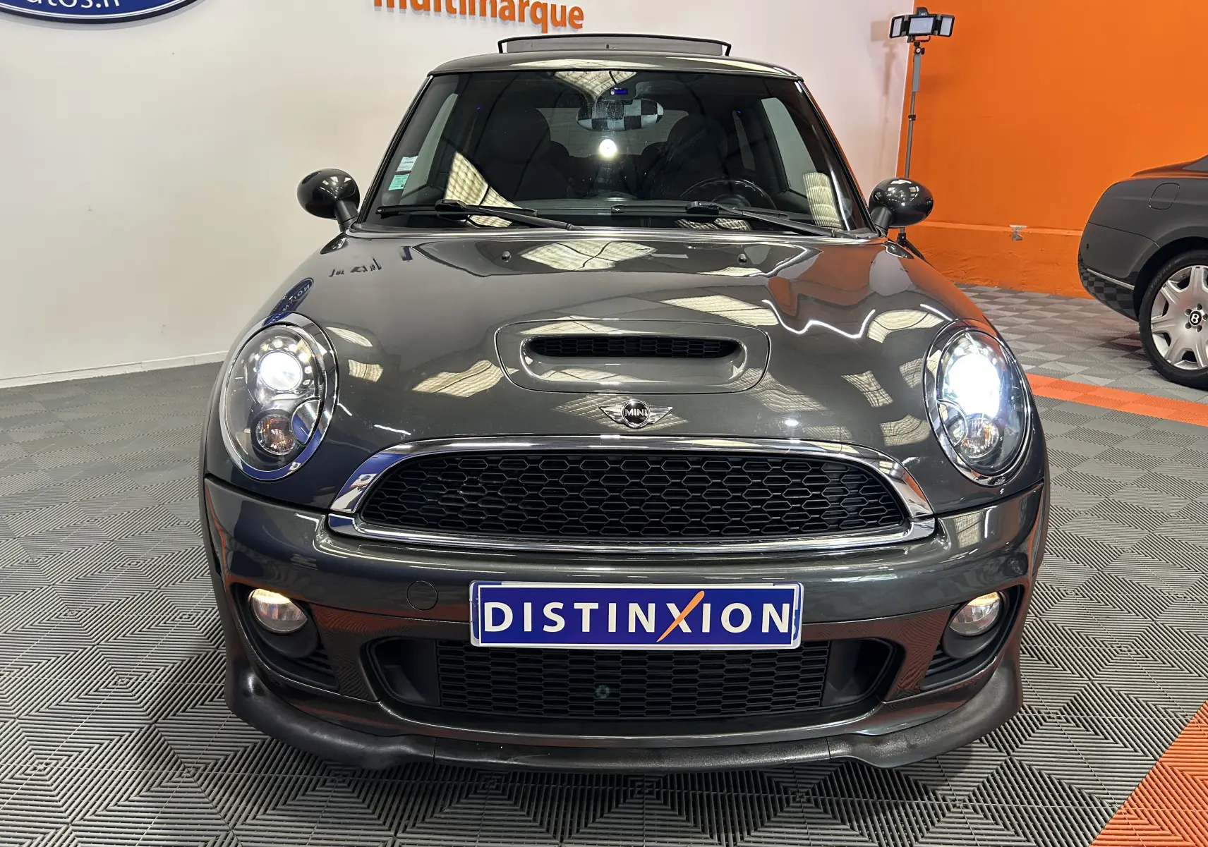 Vue avant d'une MINI Cooper gris foncé avec phares allumés, calandre noire et plaque Distinxion visible.