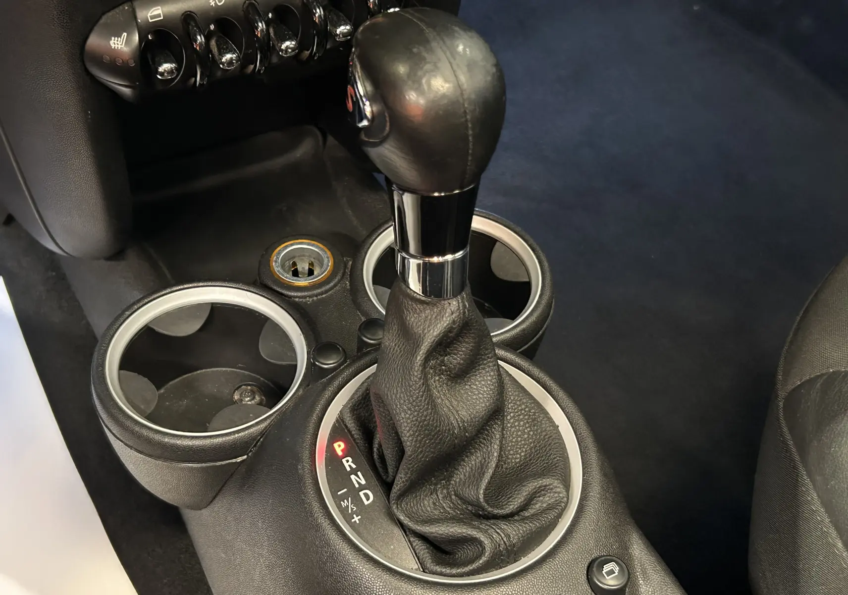 Gros plan sur le levier de vitesses automatique noir de la MINI Cooper gris, avec porte-gobelets et commandes en arrière-plan.