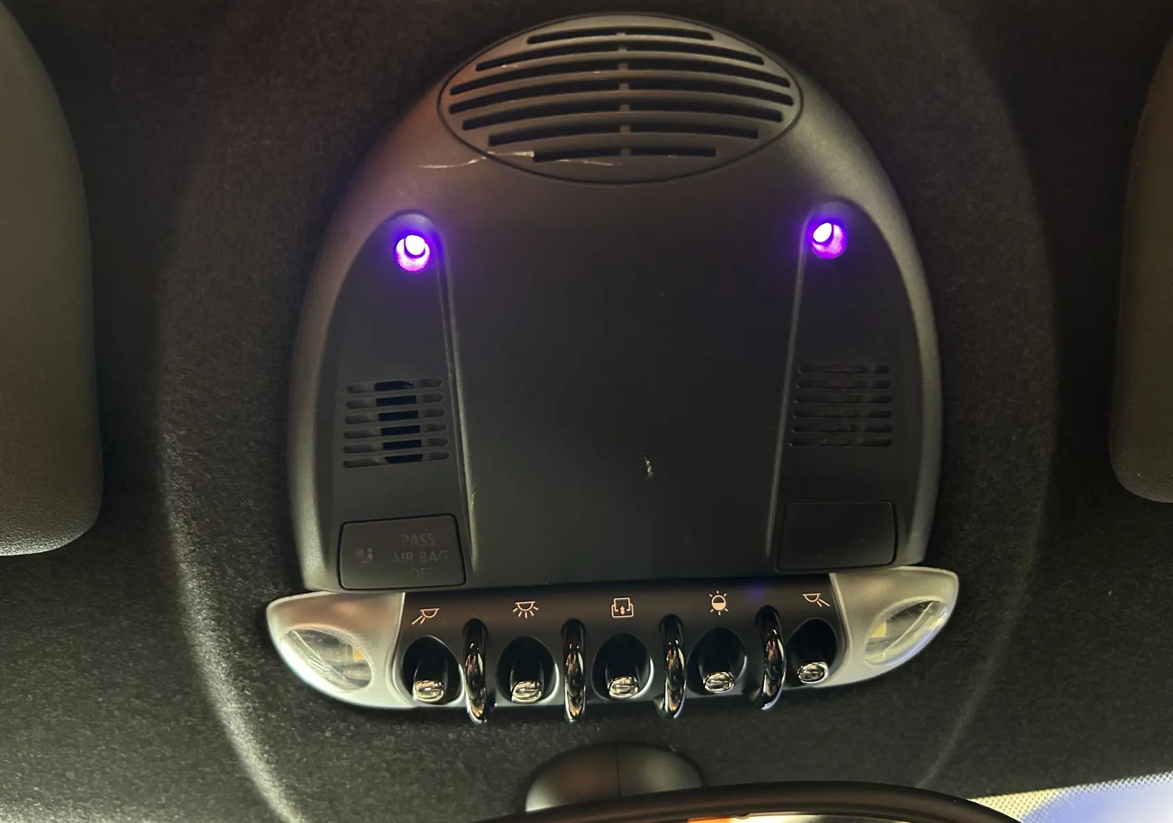 Vue en plongée sur le plafonnier noir de la MINI Cooper gris 2011 avec interrupteurs chromés et lumières violettes allumées.