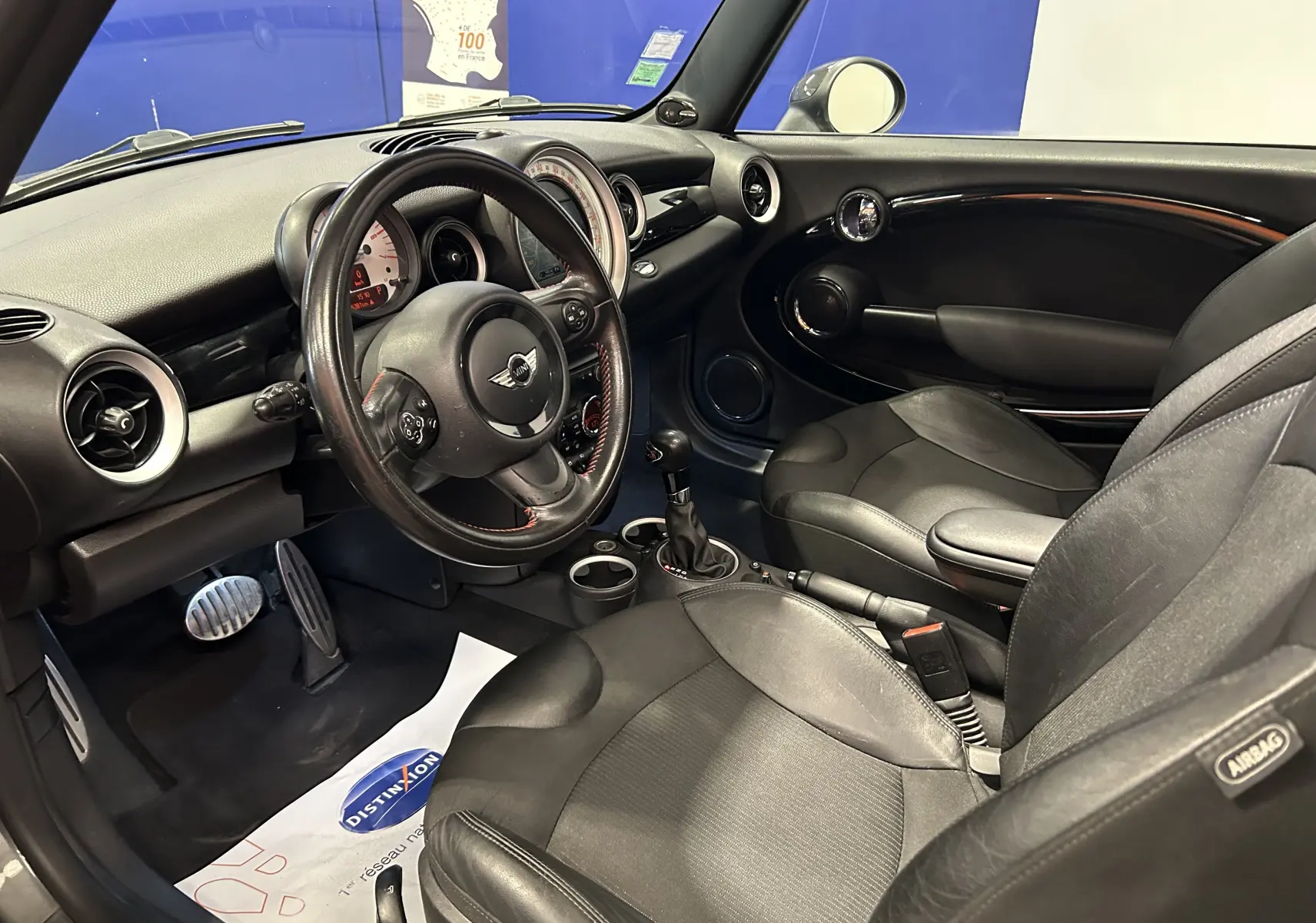 Vue intérieure côté conducteur de la MINI Cooper gris, avec volant cuir, sièges noirs et tableau de bord rond caractéristique.