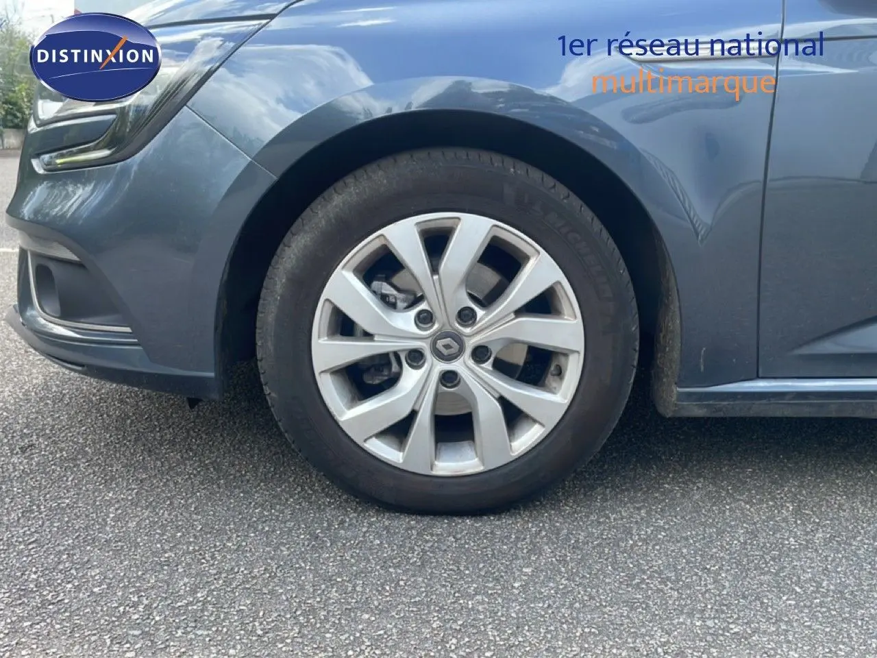 Gros plan sur la roue avant gauche d'une Renault Mégane gris titanium métal, avec jante alliage et partie avant du véhicule visible.