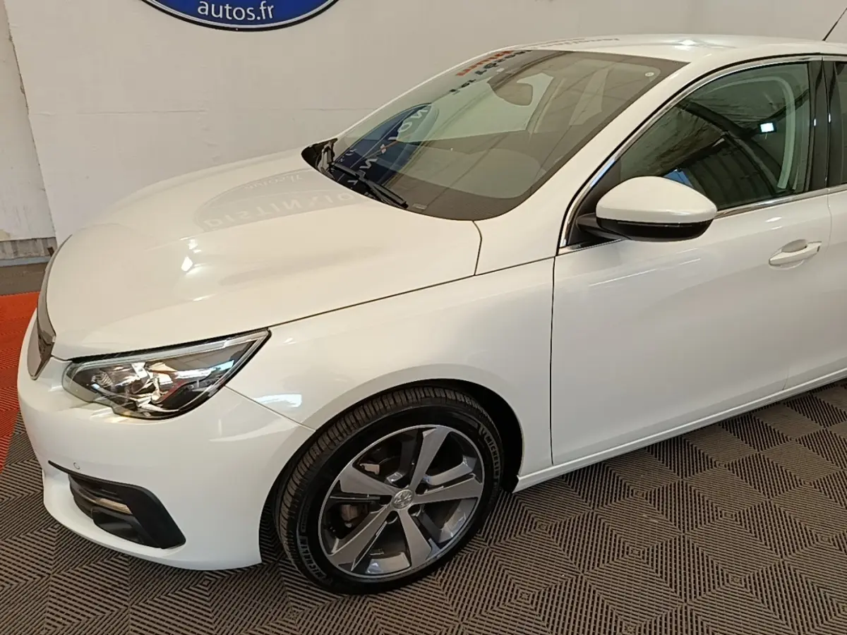 Vue 3/4 avant droit d'une Peugeot 308 blanche avec jantes alliage et feux avant LED visibles en intérieur showroom.