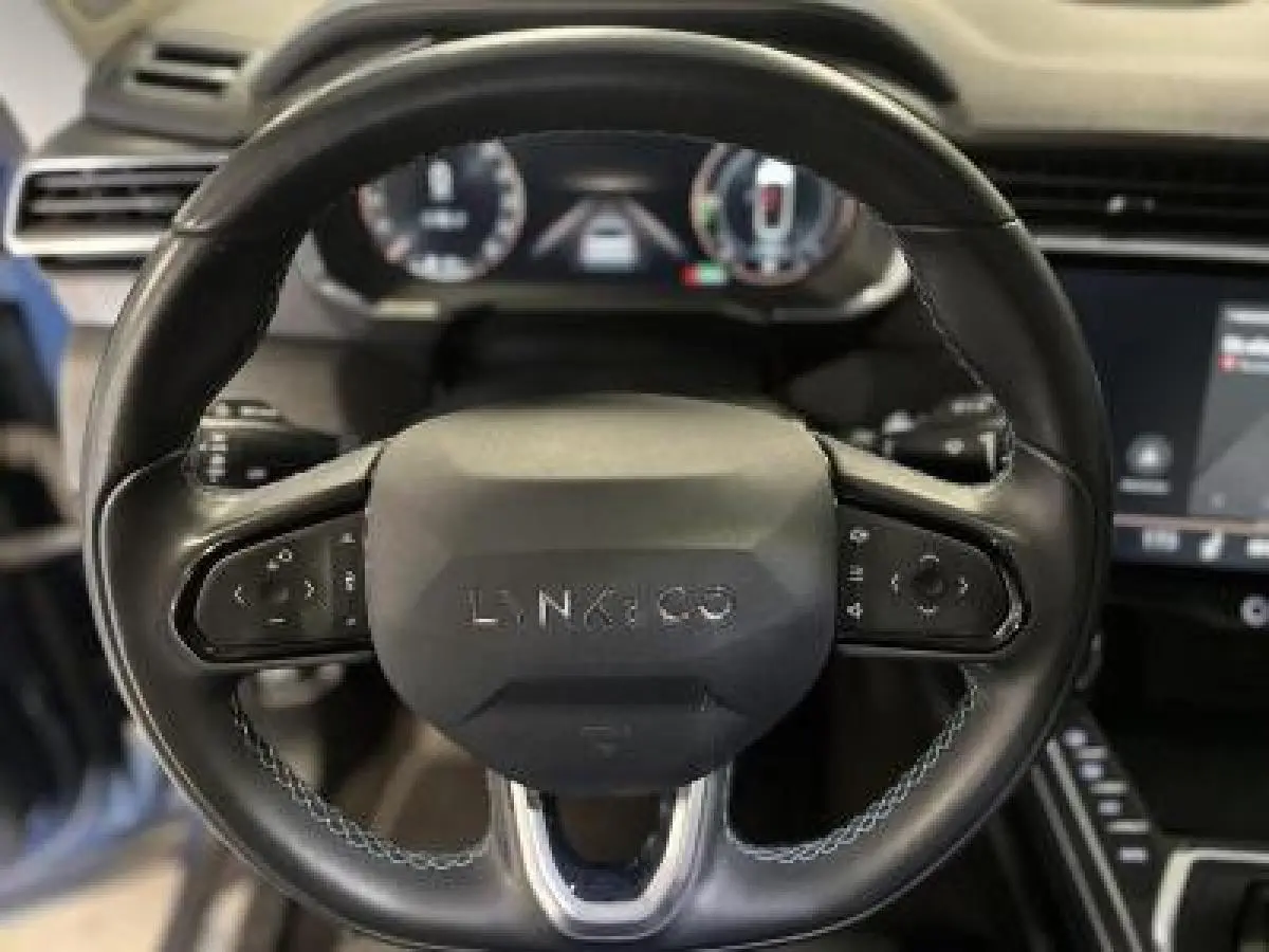 Vue rapprochée du volant noir du Lynk & Co 01 1.5 197 Hybride avec commandes intégrées et tableau de bord numérique flou.