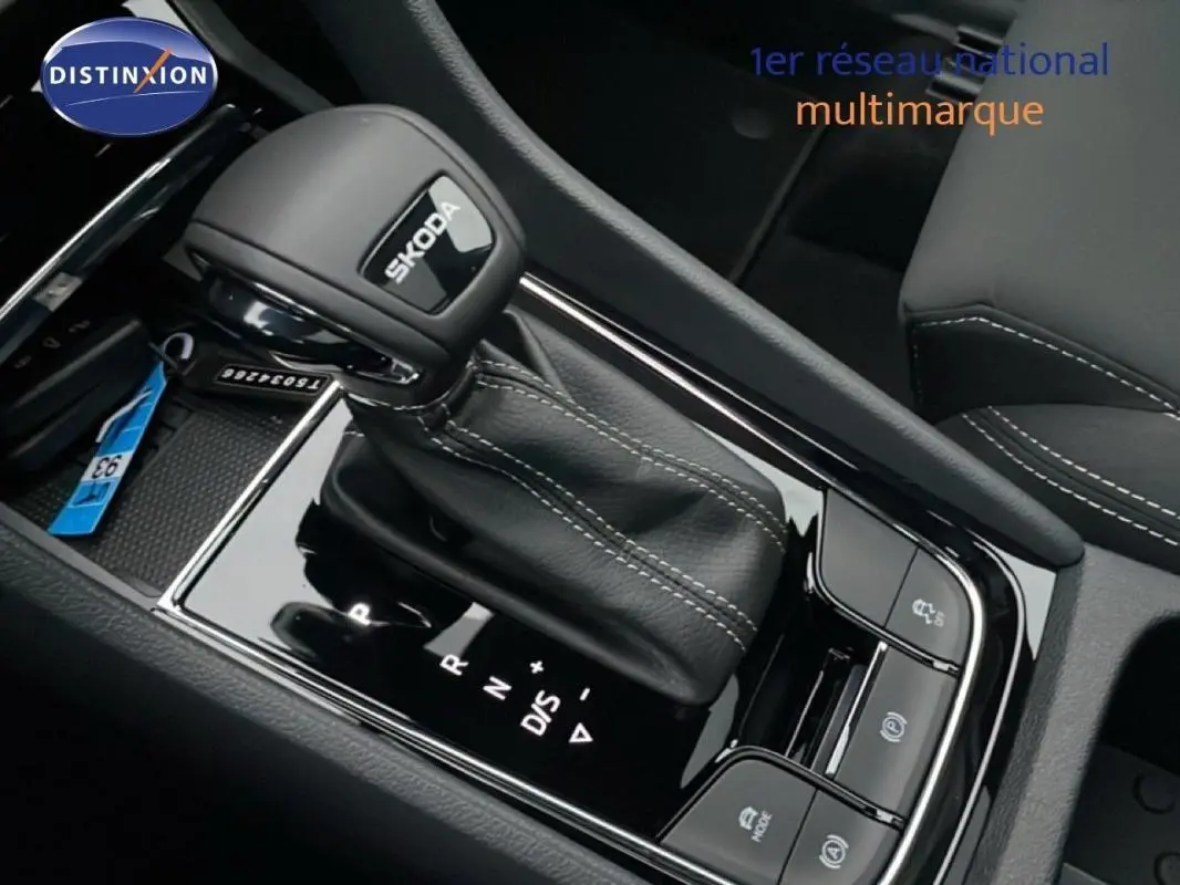 Gros plan sur la commande de boîte automatique noire avec logo Skoda, entourée de boutons de contrôle dans un intérieur sombre.