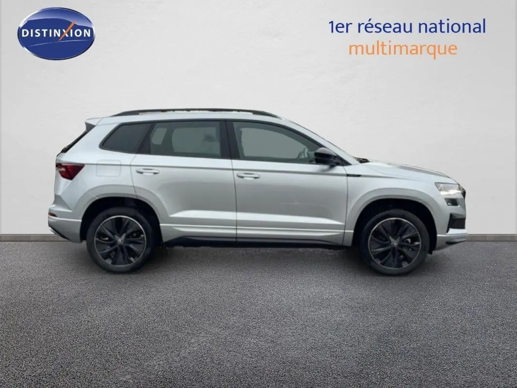 Vue latérale droite d'un Skoda Karoq argent 2025 avec barres de toit et jantes noires sportline.