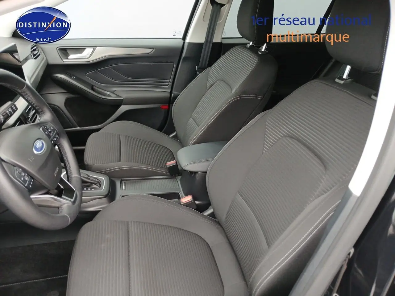 Vue intérieure côté conducteur de la Ford Focus 2024, sièges tissu noir avec accoudoir central et volant cuir multifonction.