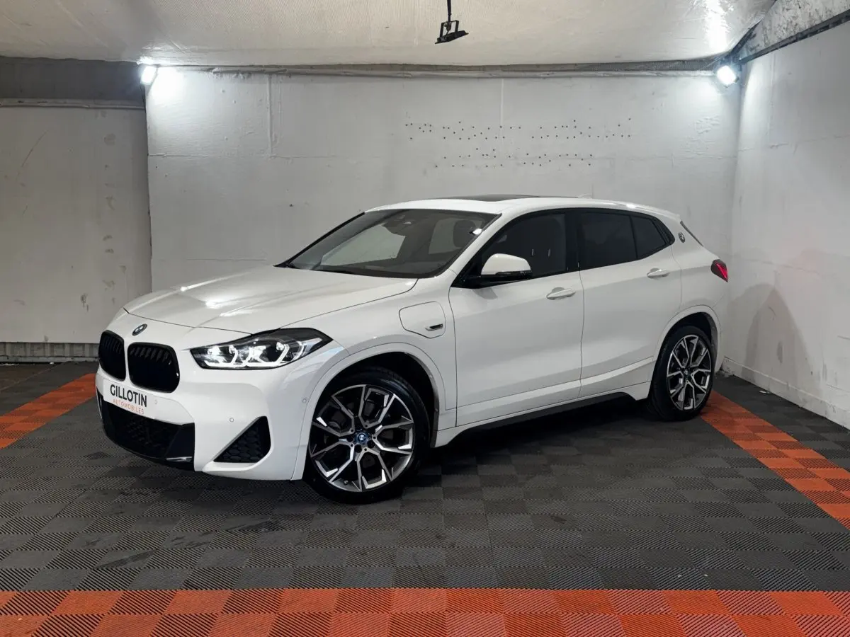 BMW X2 xDrive25e blanc en 3/4 avant droit, avec jantes alliage et toit ouvrant panoramique visible.