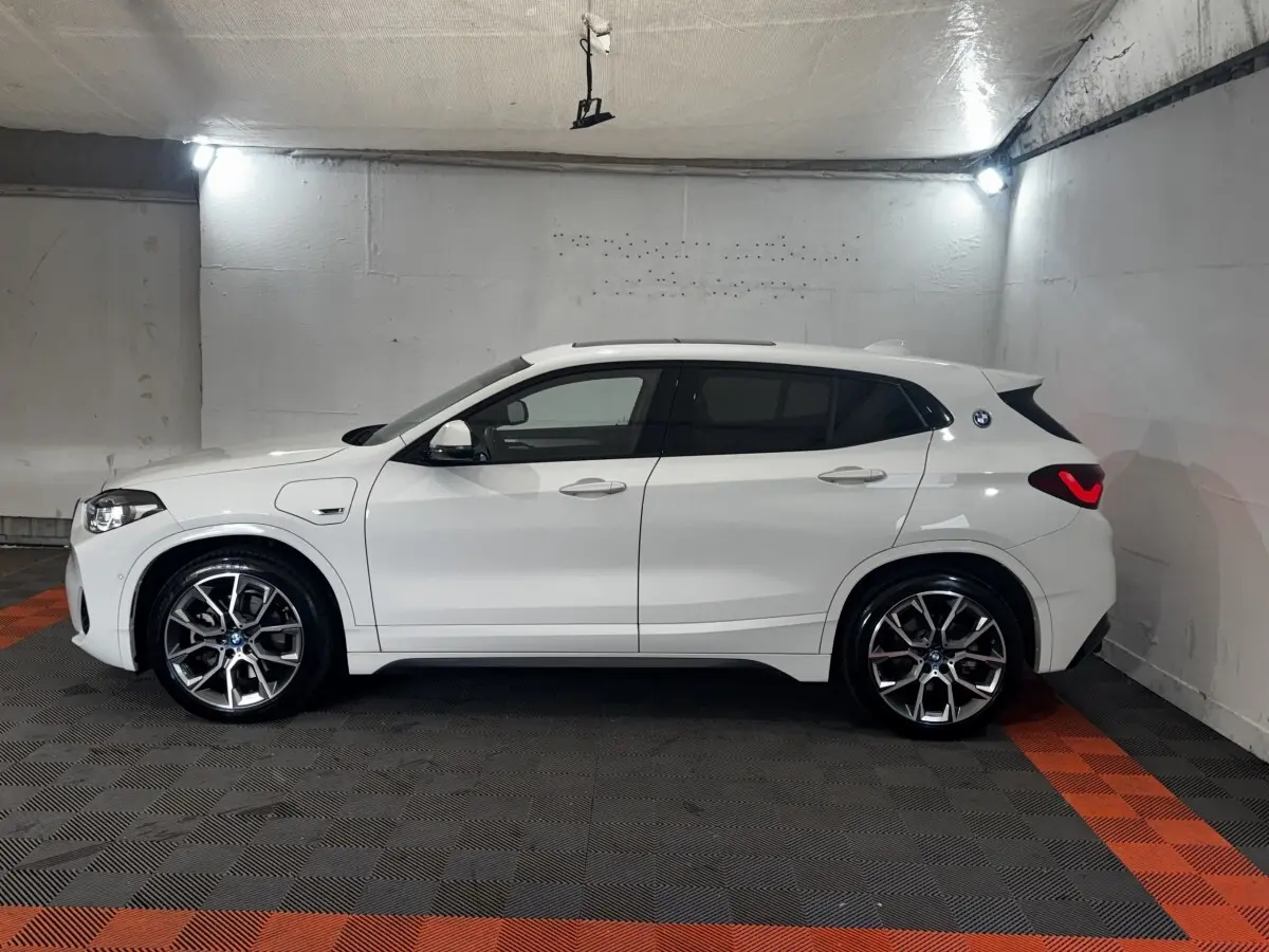 BMW X2 xDrive25e blanc en vue profil côté gauche dans un garage, avec jantes alliage et logo M Sport visible.