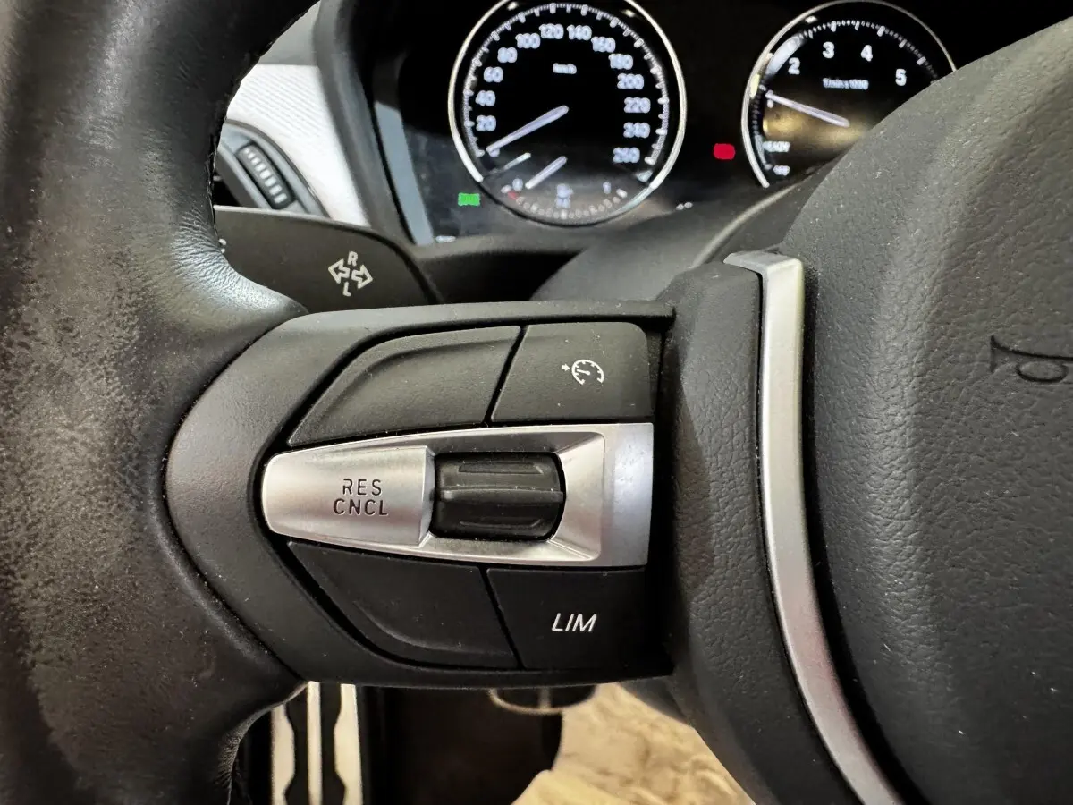 Gros plan sur les commandes de régulateur de vitesse à gauche du volant cuir noir d'une BMW X2 blanche, tableau de bord en arrière-plan.