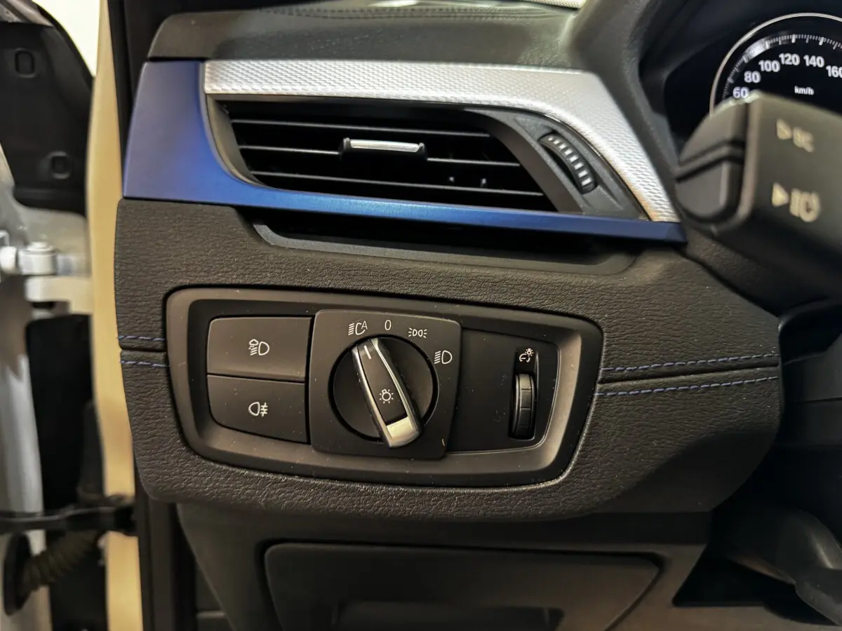 Gros plan sur le panneau de commande des phares à gauche du volant dans l’habitacle noir et bleu d’une BMW X2 blanche.