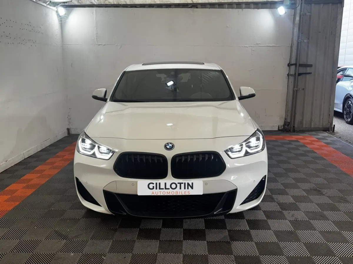 BMW X2 xDrive25e blanc vue de face avec phares LED allumés et calandre noire sportive dans un garage.