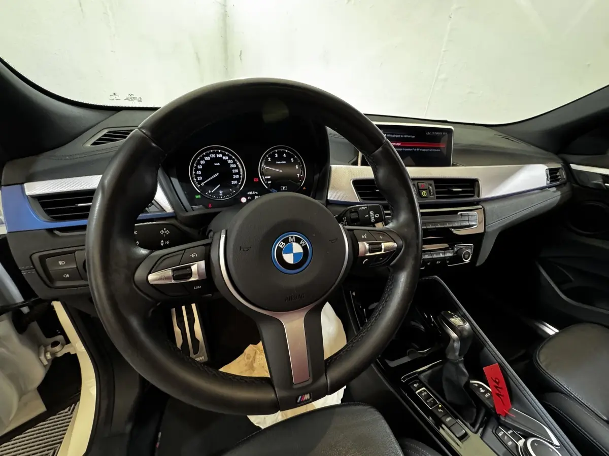 Vue intérieure centrée sur le volant cuir M et tableau de bord avec écran couleur de la BMW X2 blanc hybride.