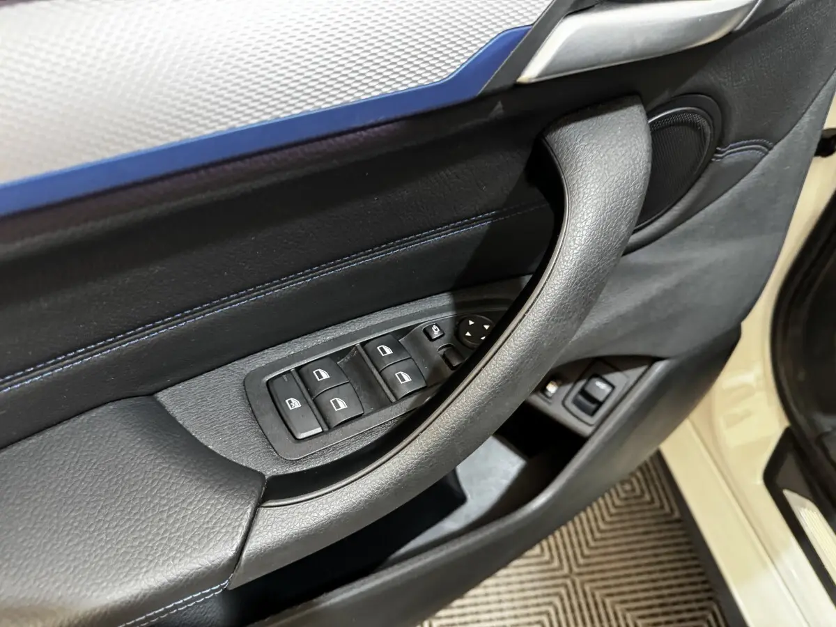 Gros plan sur la porte avant gauche noire avec commandes des vitres et poignée, BMW X2 blanc visible en arrière-plan.