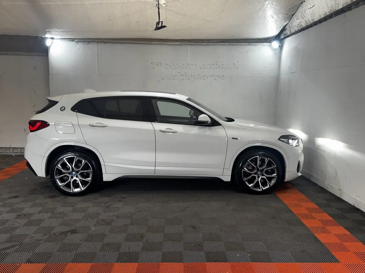 Vue de profil côté gauche d’un BMW X2 blanc avec jantes alliage et feux arrière LED allumés dans un parking fermé.