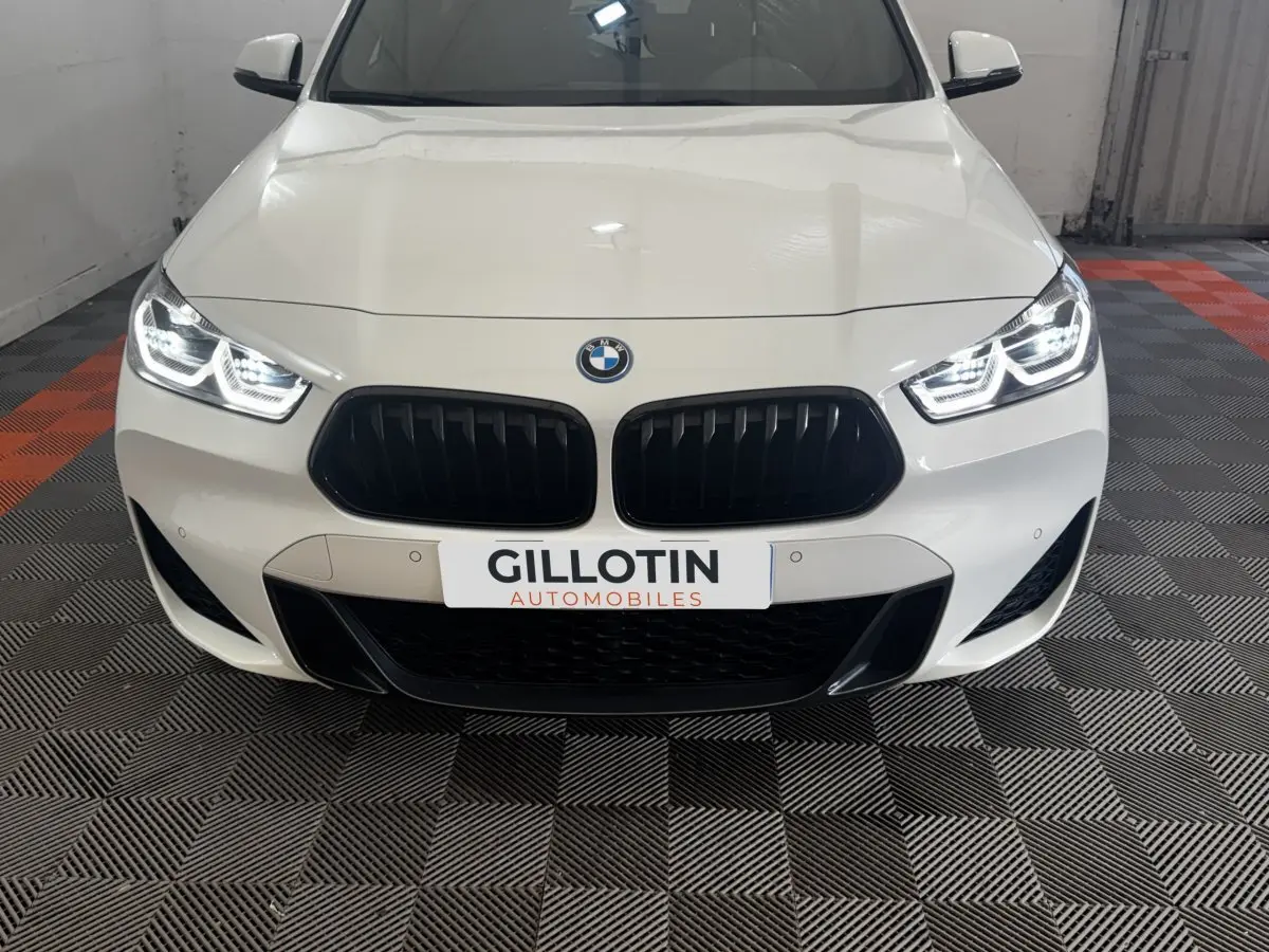 Vue avant d’une BMW X2 xDrive25e blanche avec calandre noire et phares LED allumés dans un garage.