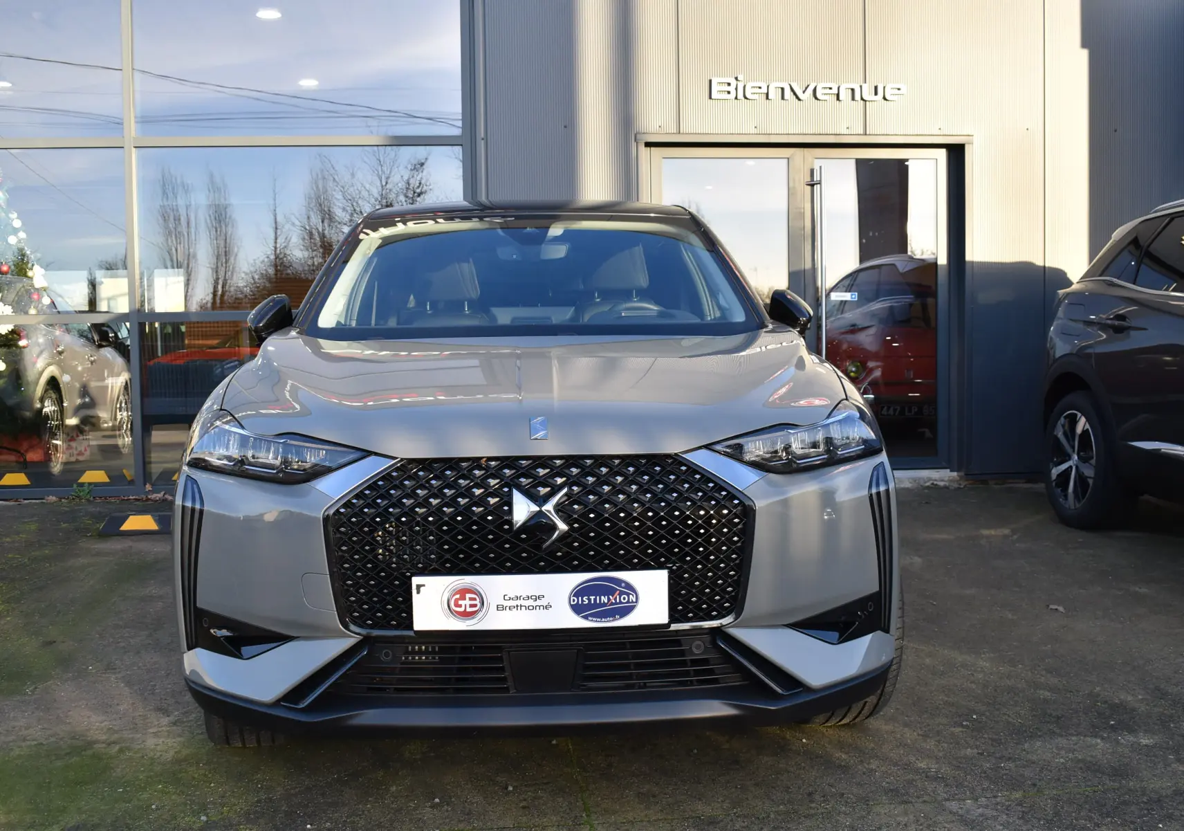 Vue avant d'une DS3 Crossback 2024 gris métallisé avec calandre noire à motif losange, devant un bâtiment avec porte vitrée.