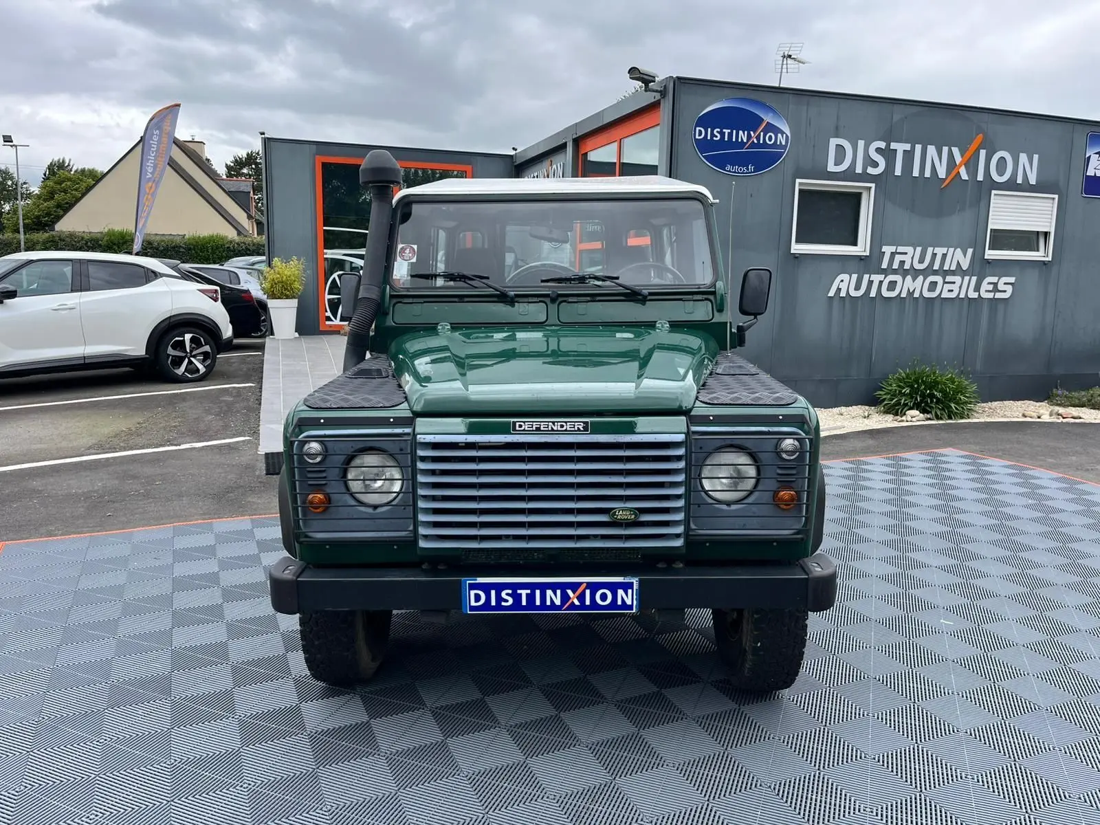 Vue frontale d'un Land Rover Defender 90 vert avec snorkel et marchepieds, stationné devant un concessionnaire.