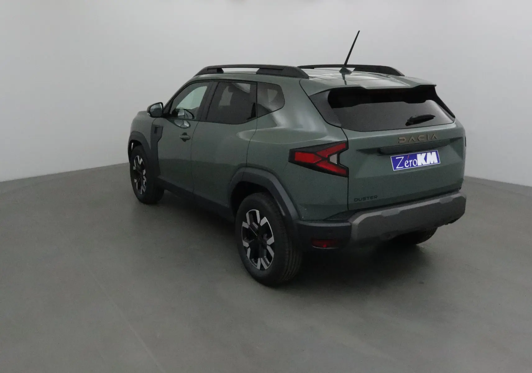 Vue 3/4 arrière gauche d'un Dacia Duster 2025 kaki lichen avec barres de toit et feux arrière en forme de flèche.