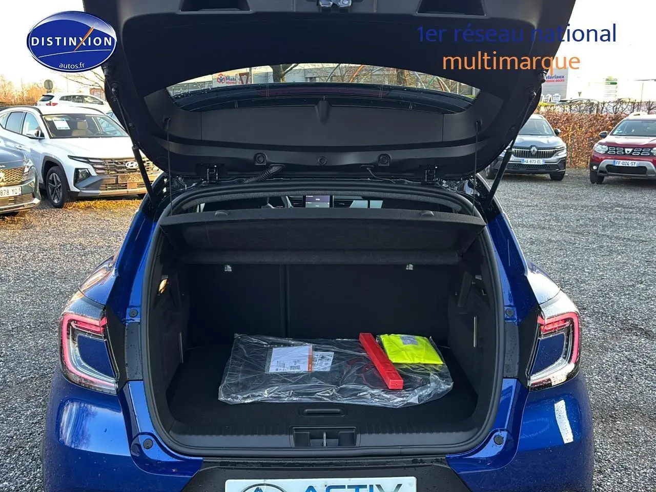 Vue arrière d’un Renault Captur bleu iron avec coffre ouvert montrant un triangle de signalisation et un gilet jaune à l’intérieur.