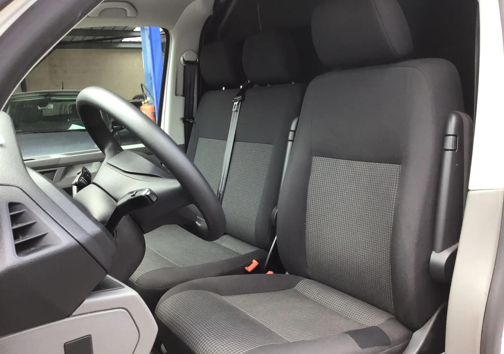Vue intérieure côté conducteur du Volkswagen Transporter 6.1 Van blanc, montrant les sièges avant en tissu gris et noir.