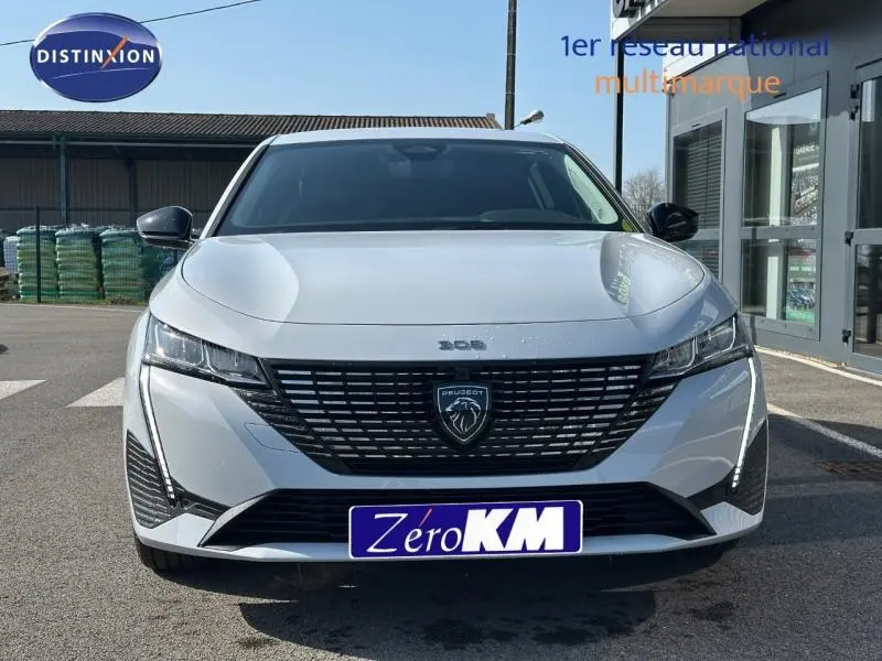 Vue de face d'une Peugeot 308 blanche avec calandre noire et logo lion central, stationnée devant un bâtiment.