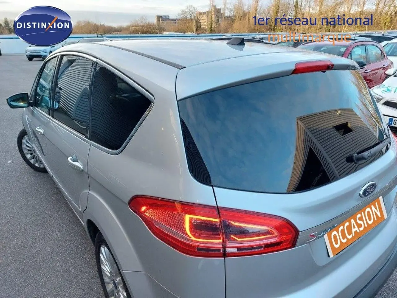 Vue 3/4 arrière droite d'un Ford S-MAX gris clair métal avec feux arrière allumés sur parking extérieur.
