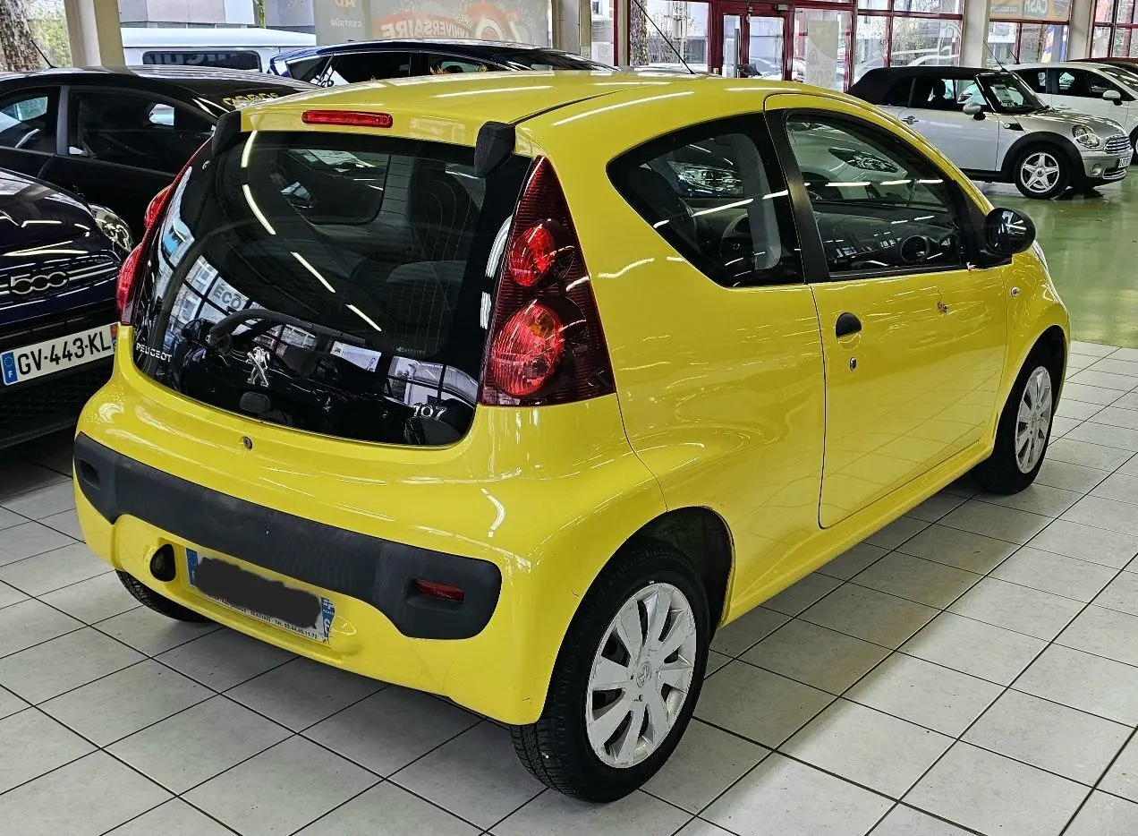 Vue 3/4 arrière droite d'une Peugeot 107 jaune vif en intérieur, avec feux arrière verticaux et pare-chocs noir.