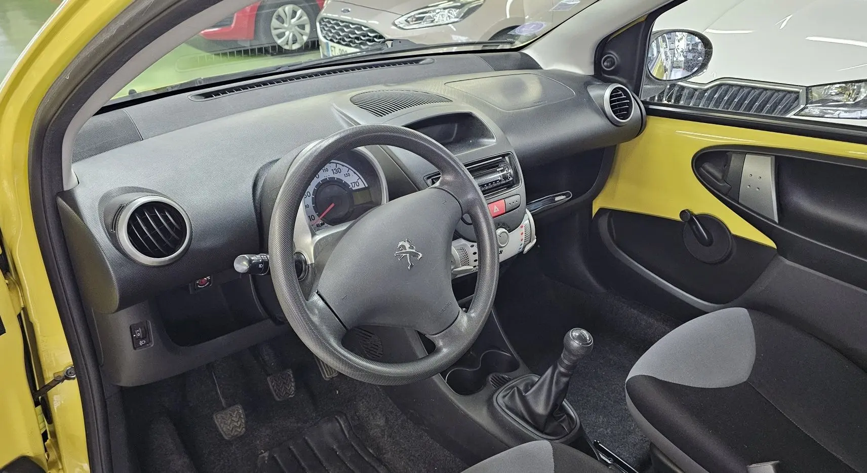 Intérieur côté conducteur d'une Peugeot 107 jaune de 2013, volant, tableau de bord et levier de vitesses manuelle visibles.