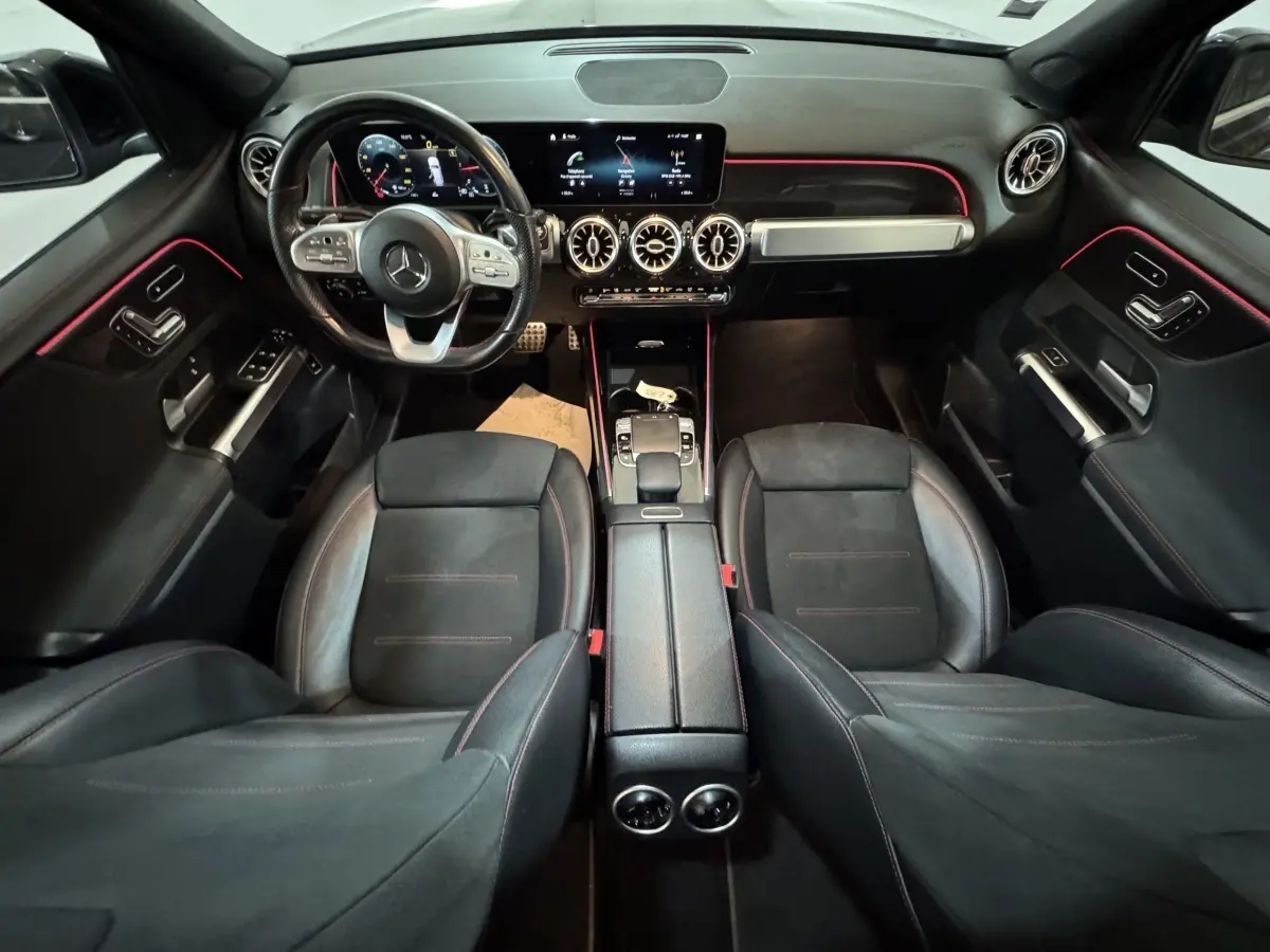 Vue intérieure avant du Mercedes GLB 200 d AMG Line noir, avec tableau de bord numérique et éclairage d'ambiance rouge.