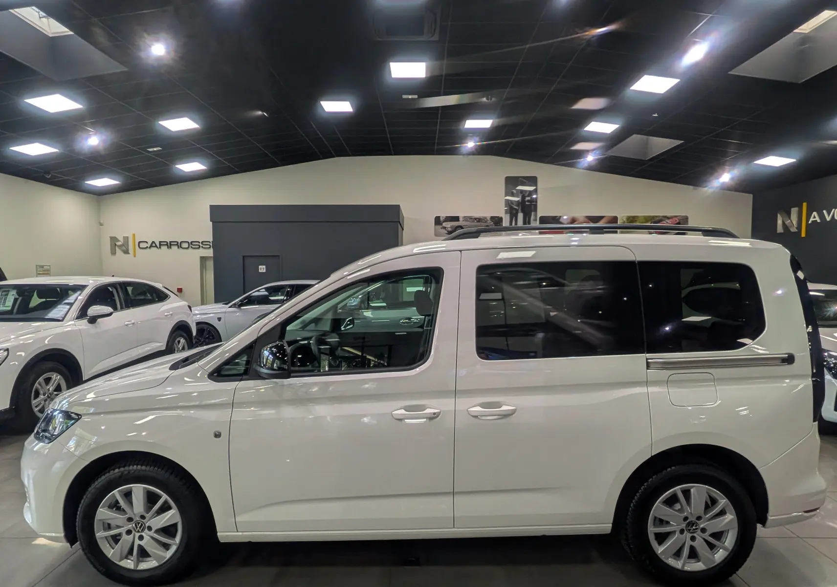 Profil latéral droit du Volkswagen Caddy blanc 2025 en showroom, avec vitres arrière teintées et jantes alliage.