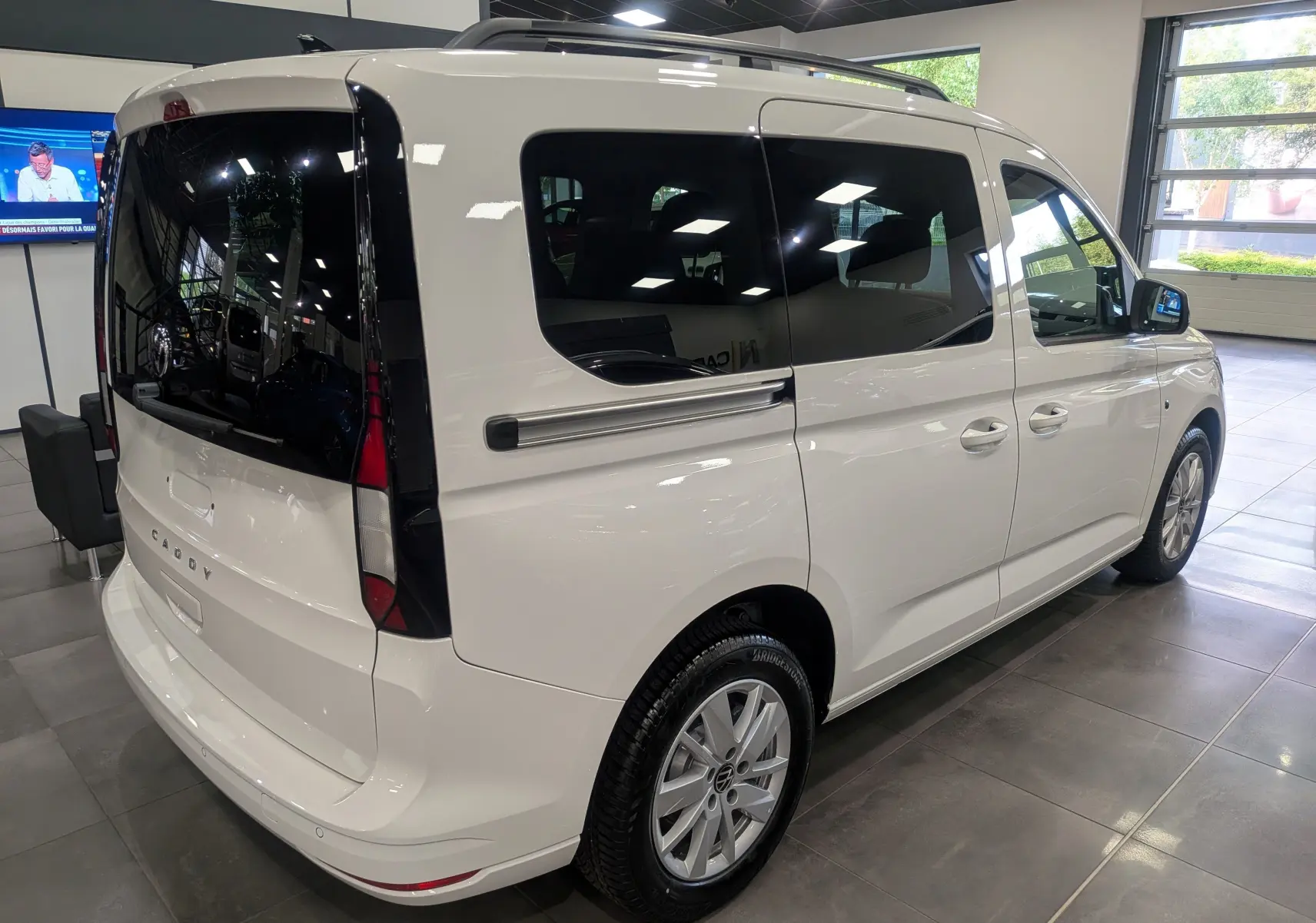 Vue 3/4 arrière droite d'un Volkswagen Caddy blanc 2025 en showroom avec vitres teintées et jantes alliage.