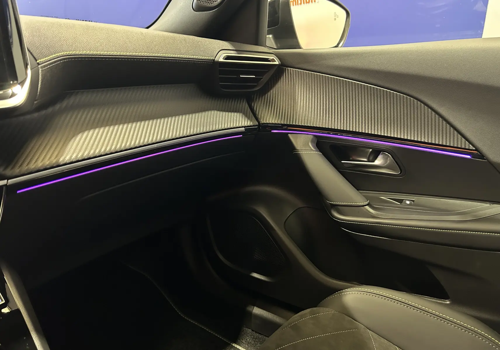 Vue intérieure côté passager du Peugeot 2008 Hybrid noir 2025, avec éclairage d'ambiance violet et finitions carbone.