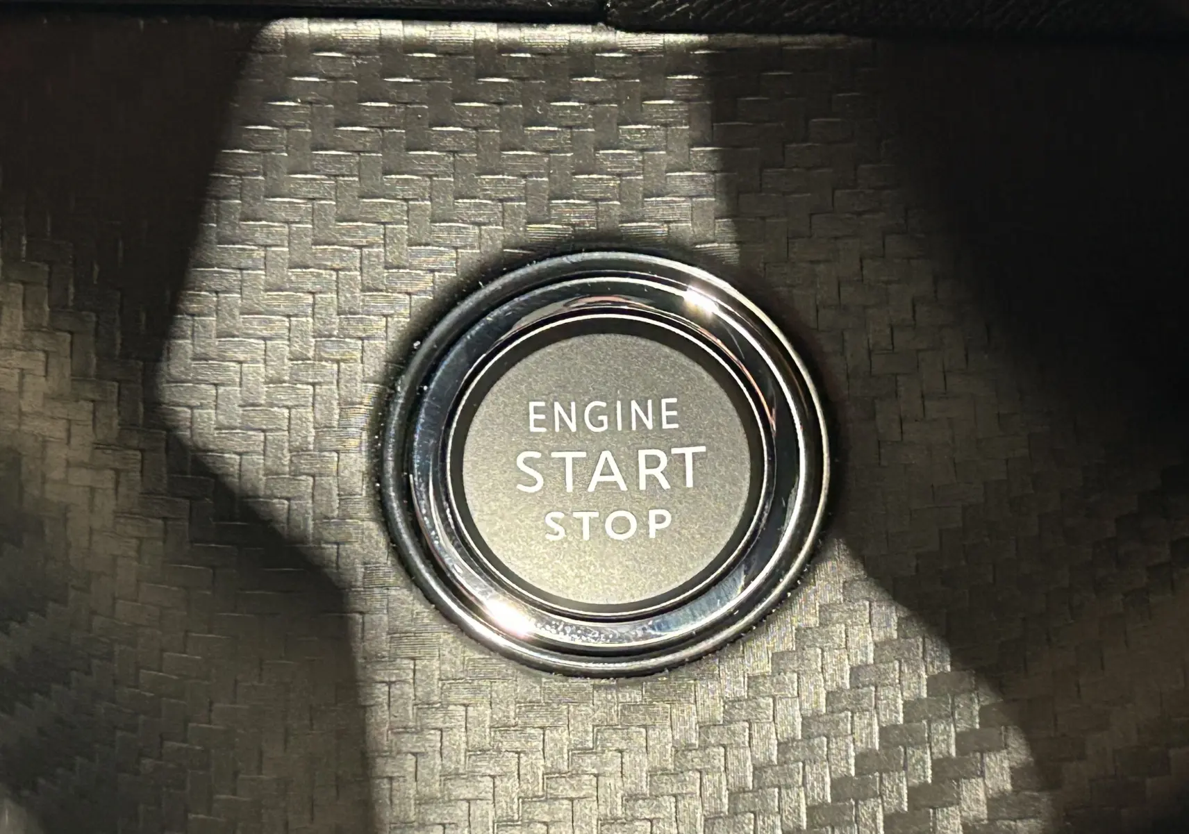 Gros plan sur le bouton moteur Start/Stop entouré d'un cerclage chromé sur un fond noir à motif carbone.