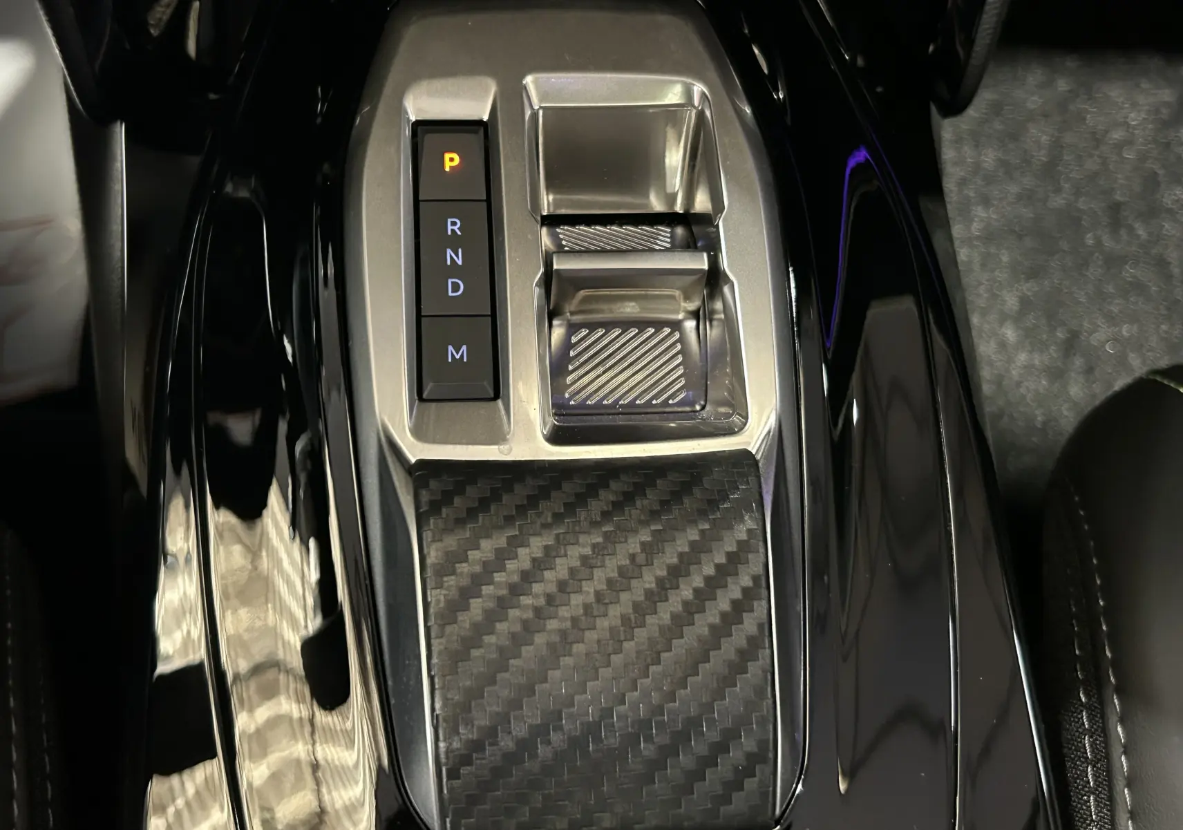 Gros plan sur la console centrale noire brillante du Peugeot 2008 Hybrid avec sélecteur de boîte automatique et finition carbone.
