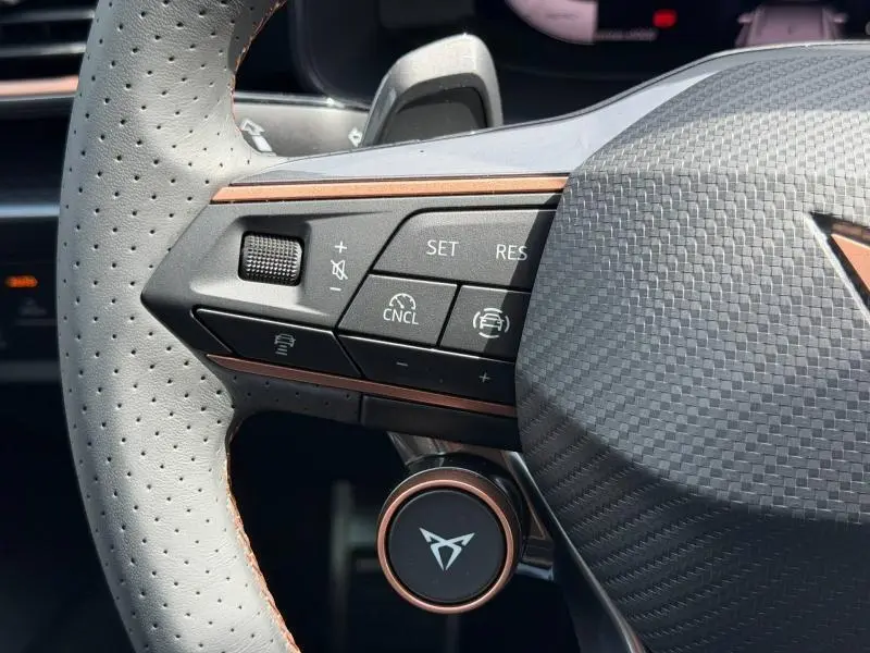 Gros plan sur le côté gauche du volant perforé gris avec commandes de régulateur et logo CUPRA sur bouton central.