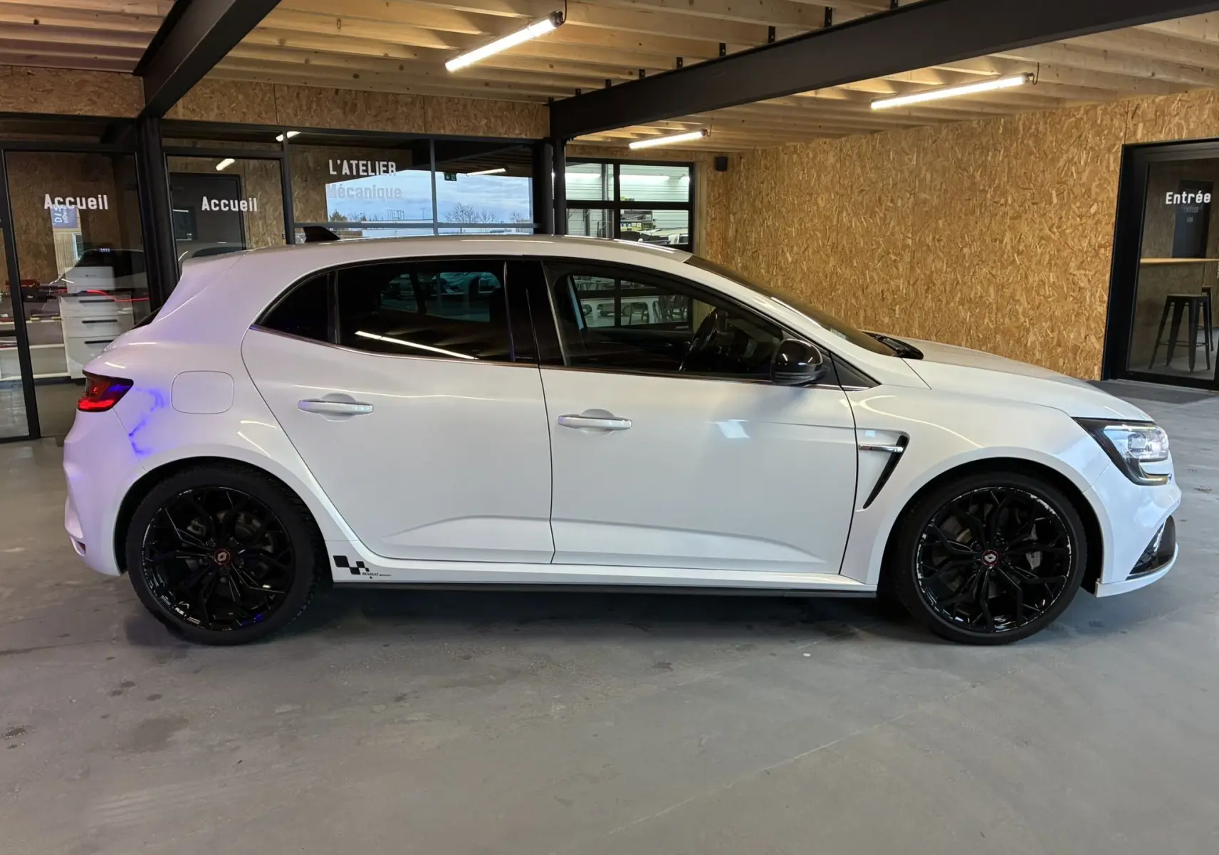 Profil droit d'une Renault Mégane IV RS 280 EDC6 blanche avec jantes noires et détails sportifs en intérieur d'atelier.