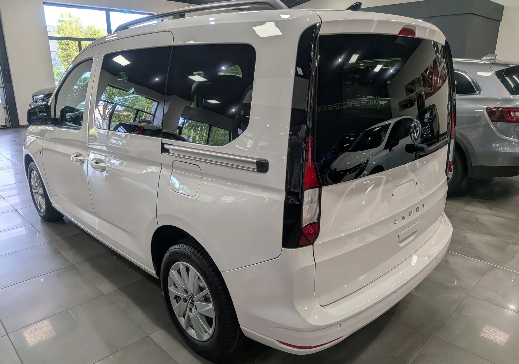 Vue 3/4 arrière droite d'un Volkswagen Caddy blanc 2025 avec vitres teintées et hayon noir brillant en showroom.