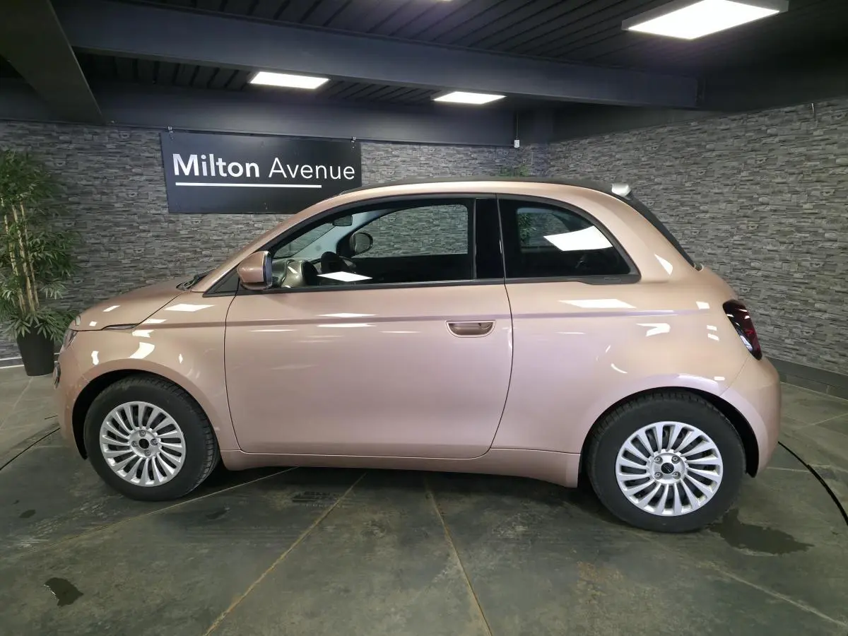Vue de profil côté gauche d'une FIAT 500 Cabriolet électrique rose métallisée avec jantes acier 15 pouces en intérieur showroom.