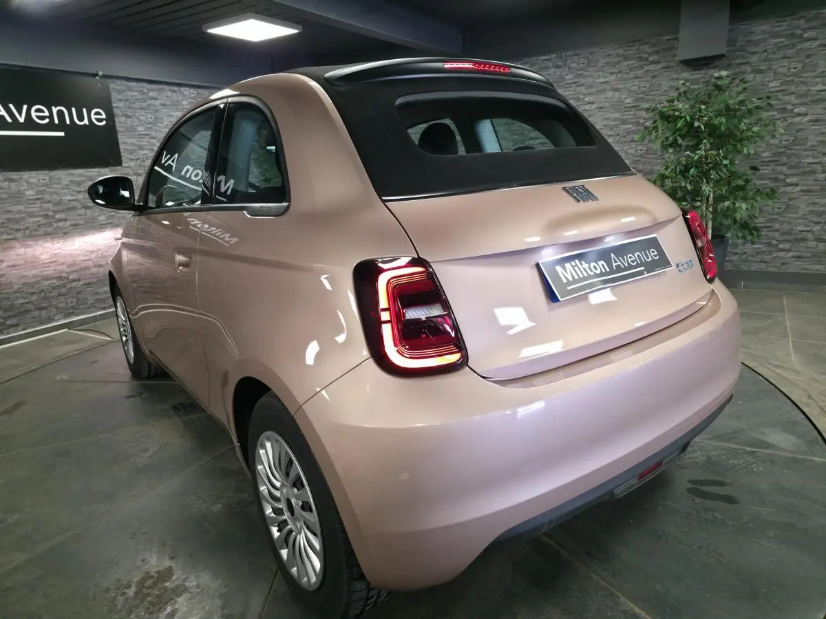 Vue 3/4 arrière droite d'une Fiat 500 Cabriolet électrique rose métallisé avec capote noire en intérieur showroom.
