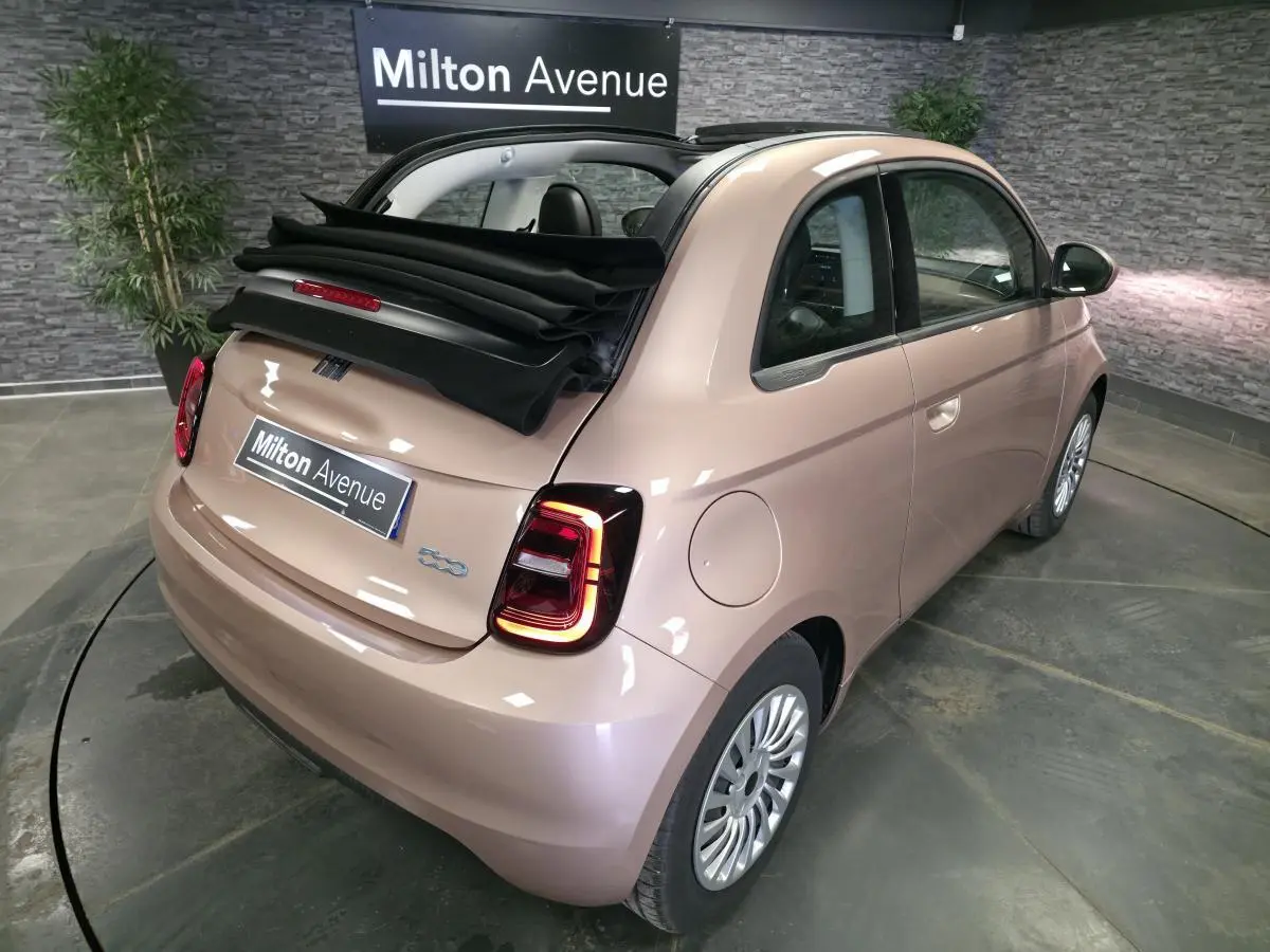 Vue 3/4 arrière droite d'une Fiat 500 Cabriolet électrique rose métallisé avec capote noire partiellement ouverte.
