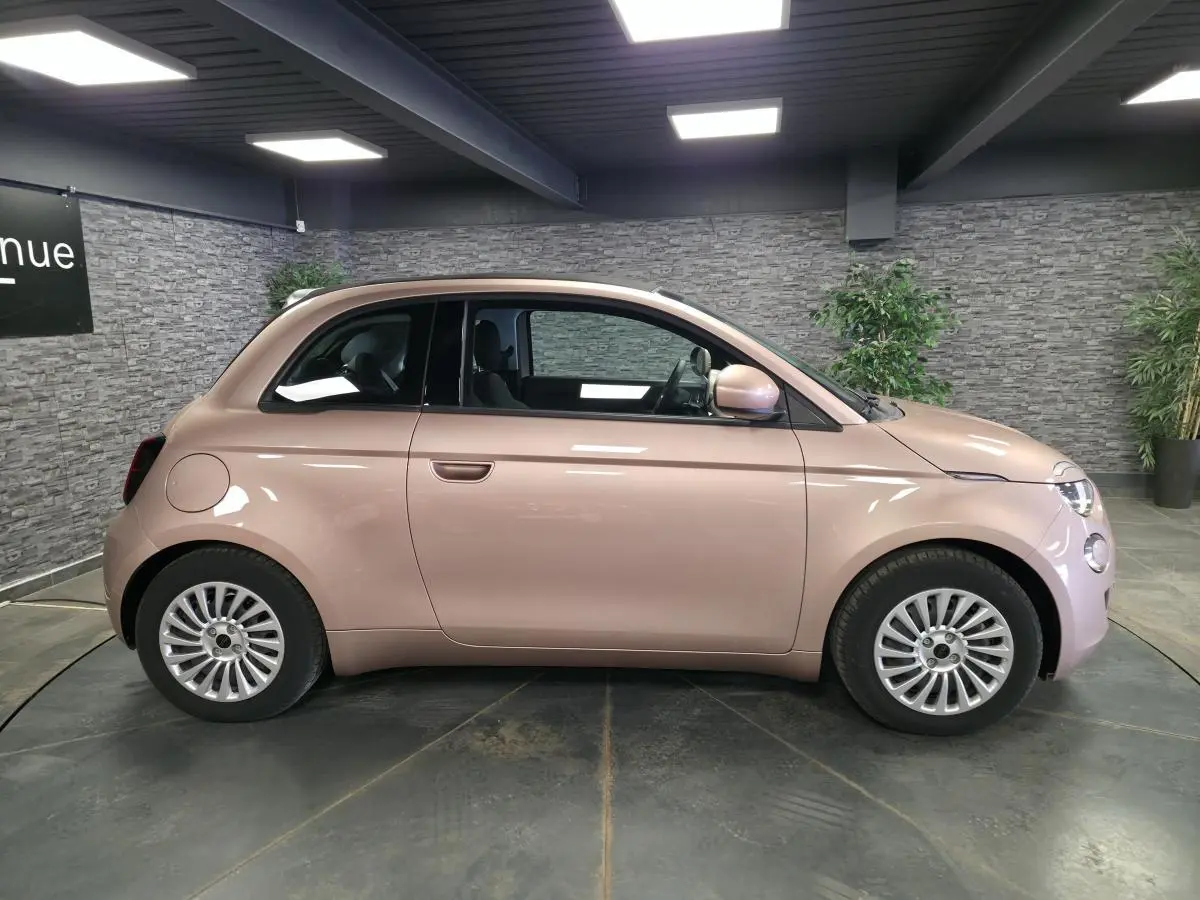 Vue de profil côté gauche d'une FIAT 500 Cabriolet électrique 2023 rose métallisée avec jantes acier et capote noire fermée.