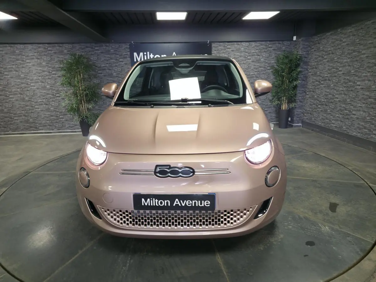 Vue avant d'une FIAT 500 Cabriolet électrique rose métallisée avec phares allumés en intérieur showroom.